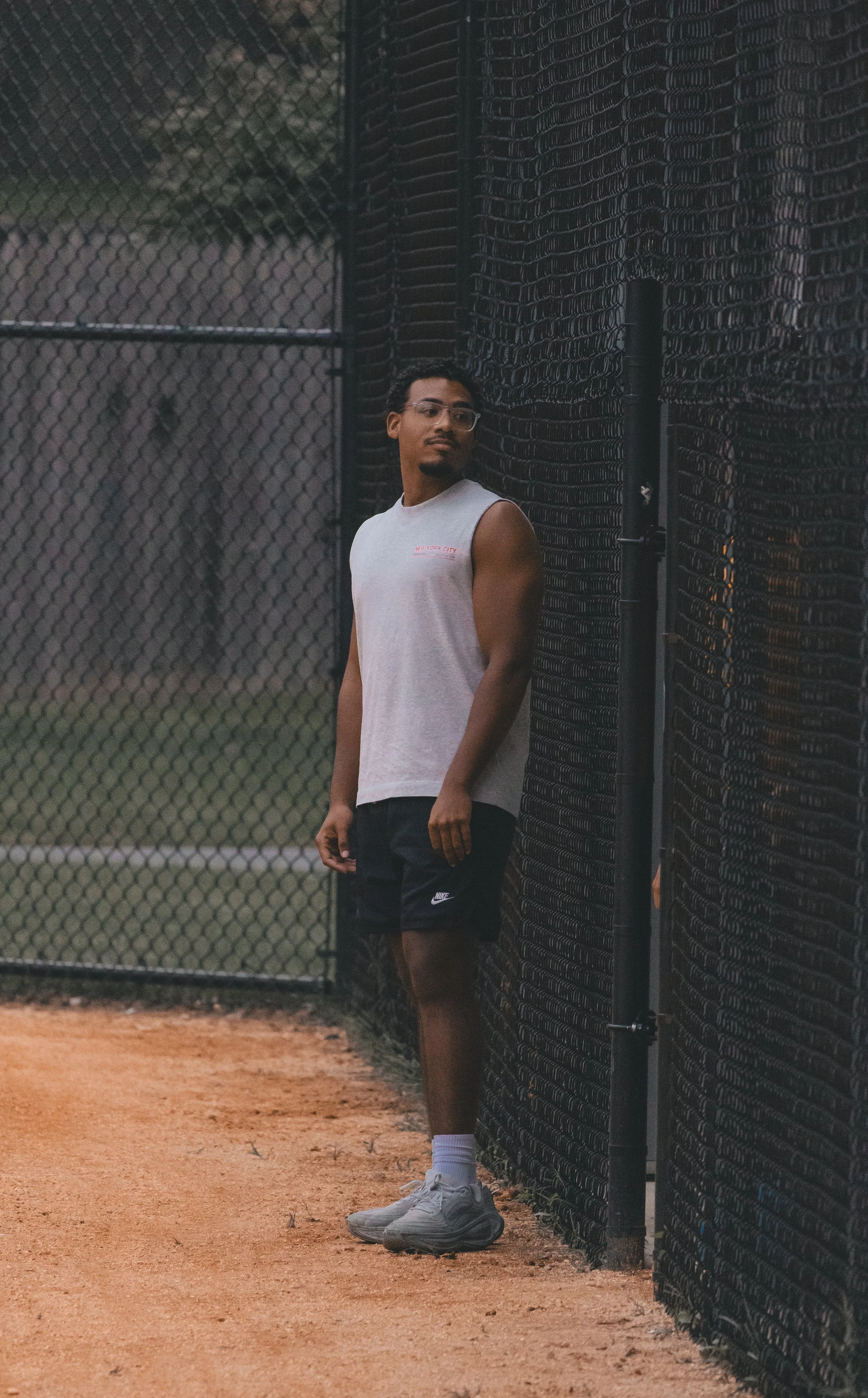 A man standing in front of a black chain-link fence on a tennis court, wearing glasses, a sleeveless white shirt, black Nike shorts, and gray tennis shoes.