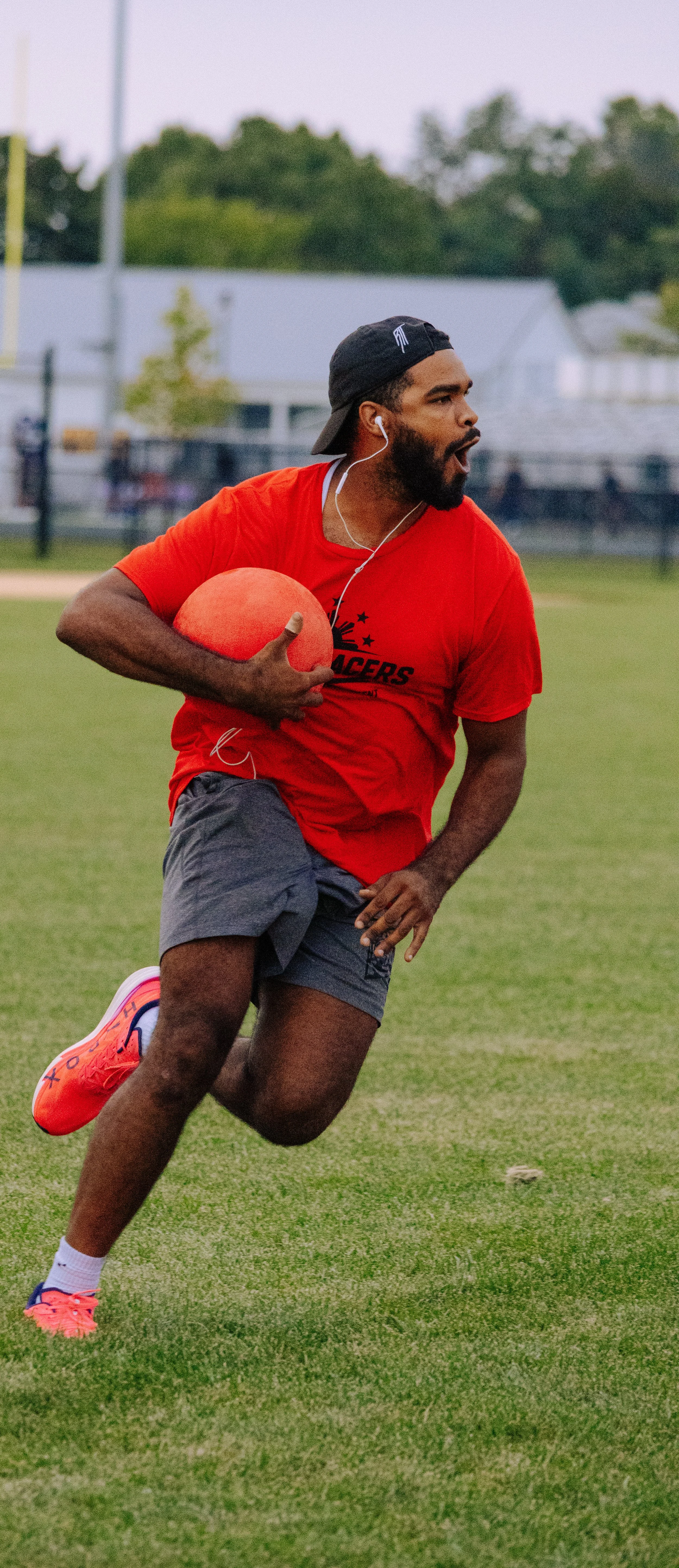 A man running on a grassy sports field holding a red medicine ball, wearing a red t-shirt, gray shorts, pink and blue athletic shoes, a black cap backward, and earphones.