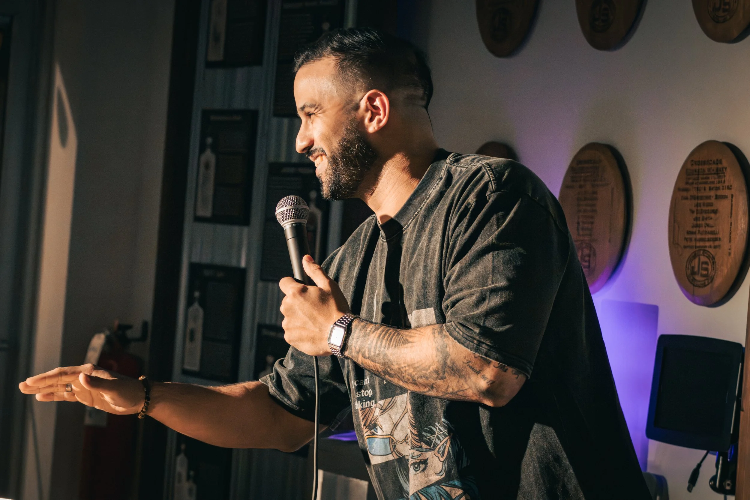 A man with tattoos on his arms and a beard, smiling and holding a microphone, appears to be speaking or performing. He is wearing a patterned shirt and a wristwatch, standing indoors against a wall decorated with wooden plaques or awards.