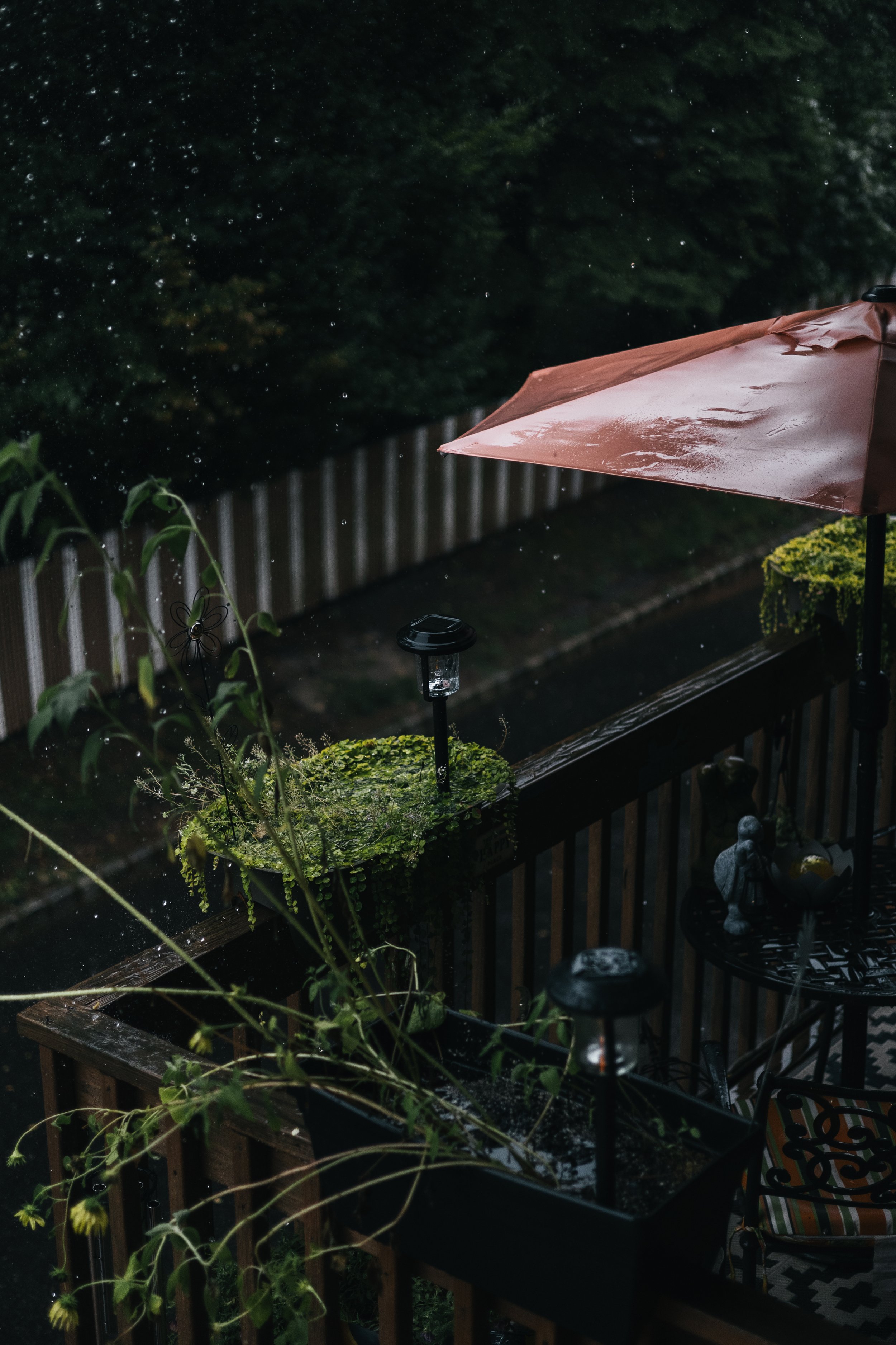 A wet patio or balcony area during a rainstorm, with a brown patio umbrella and a planter box with green plants, rain droplets visible in the air, and outdoor lights. Part of a patio table with a patterned umbrella is also visible.
