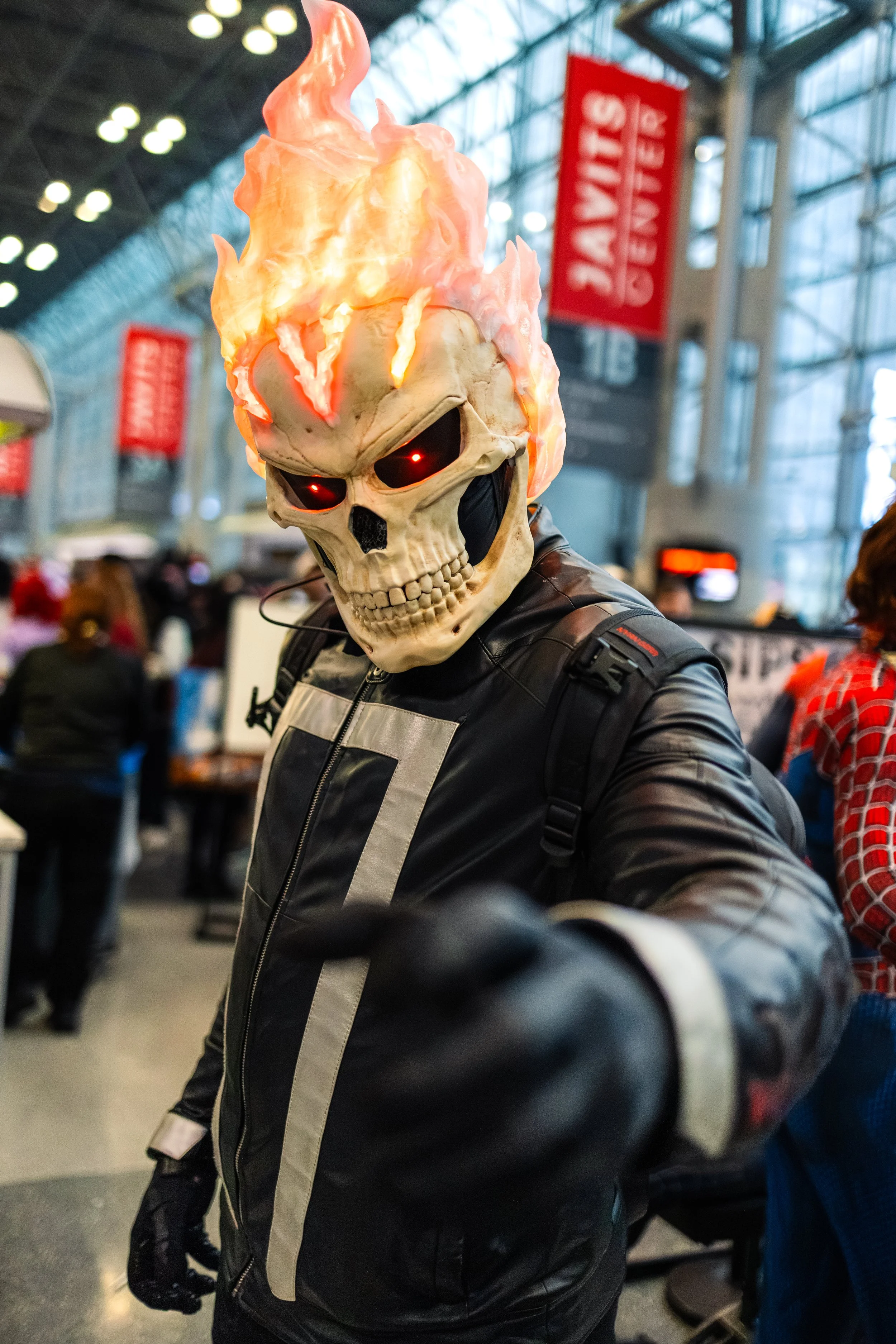 Person wearing a skull mask with flames and glowing red eyes in a busy indoor setting, likely an airport or convention center.