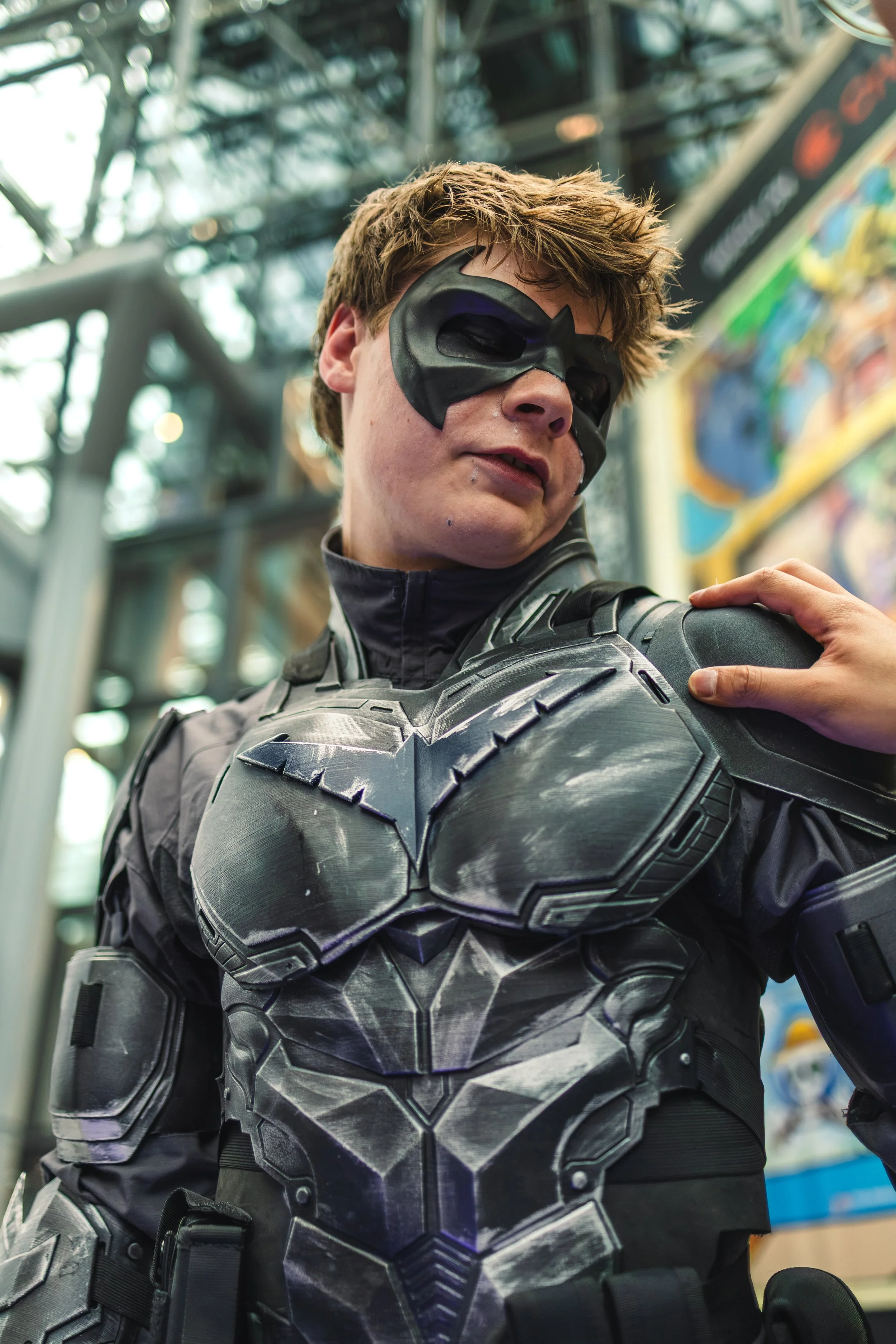 Person dressed as Batman in a black costume with a bat symbol, wearing a mask, at an indoor event or convention.