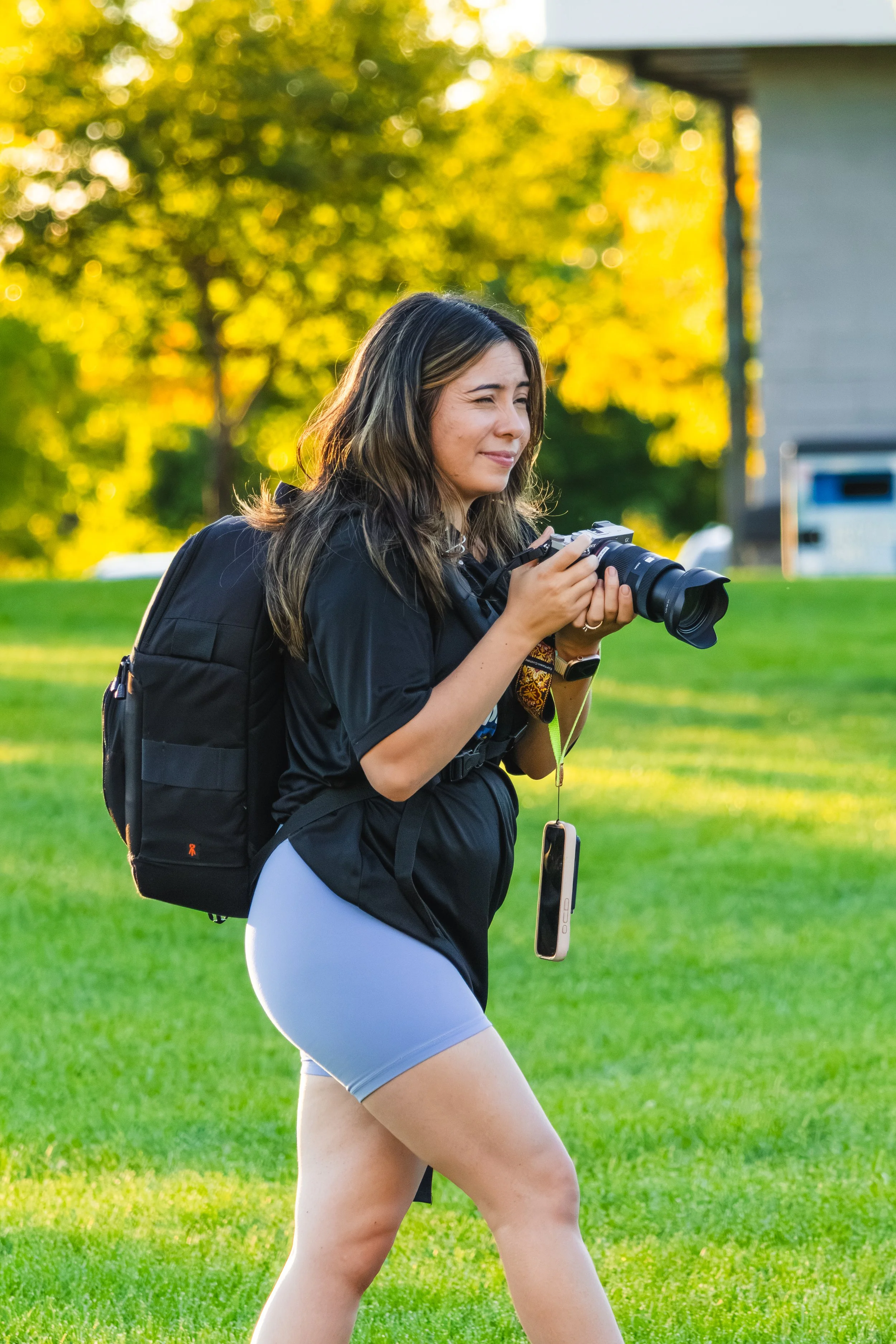 A woman walking outdoors in a park or garden on a sunny day, holding a camera, wearing a black backpack, a black jacket, and light-colored shorts, with trees and a house in the background.