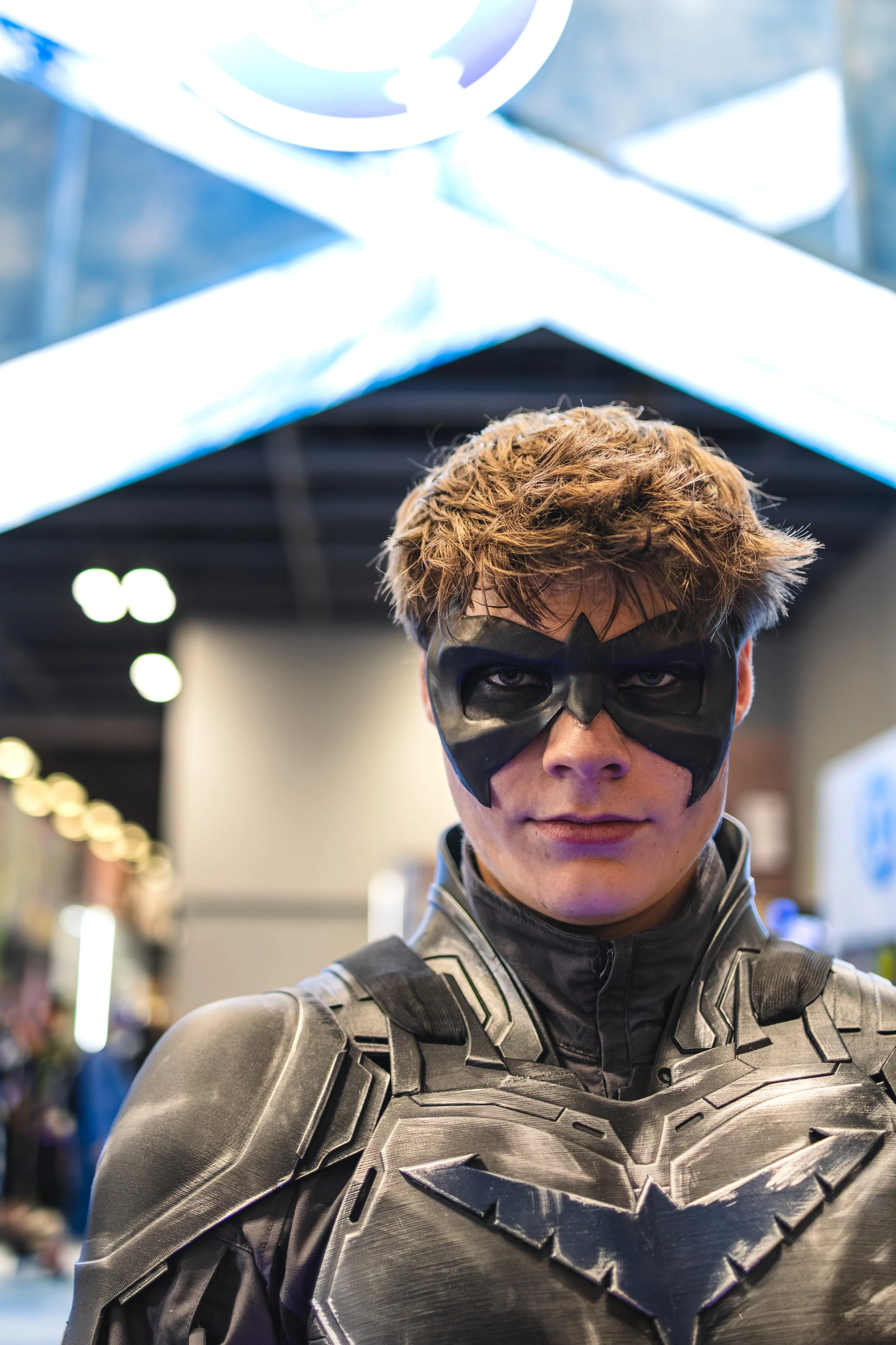 A person with a serious expression wearing a black superhero costume with metallic armor details, including a chest and shoulder piece, and a black mask covering the eyes and nose.