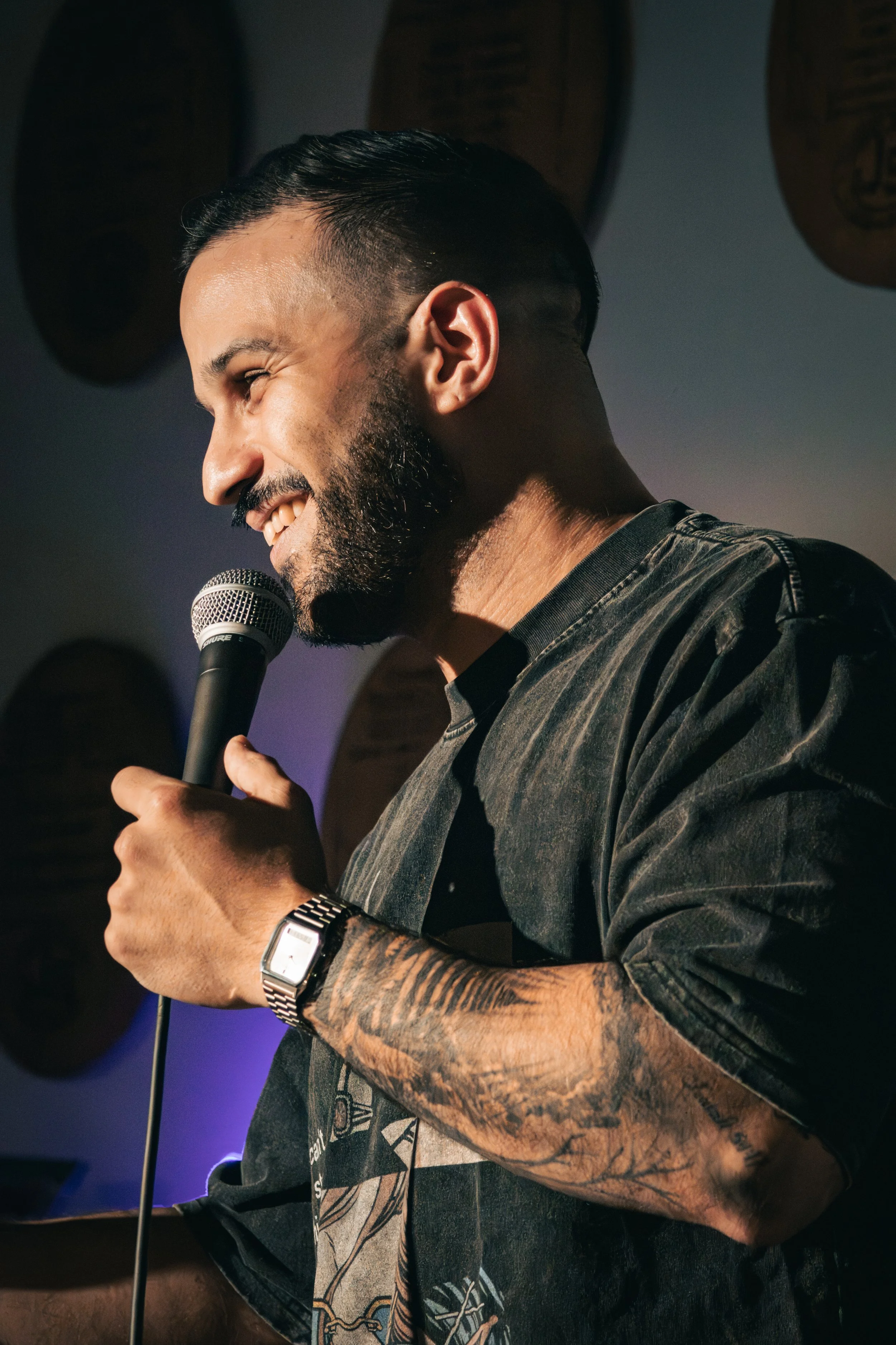 A man with a beard and a tattooed arm, smiling while holding a microphone, with drums in the background.
