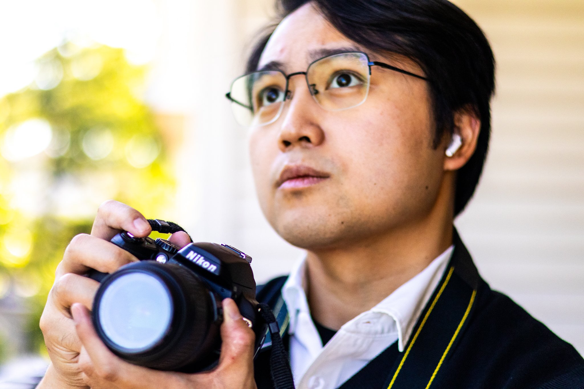 A person with short black hair, glasses, and AirPods wearing a camera strap around their neck, holding a Nikon camera with a large lens, outdoors with blurred green foliage in the background.