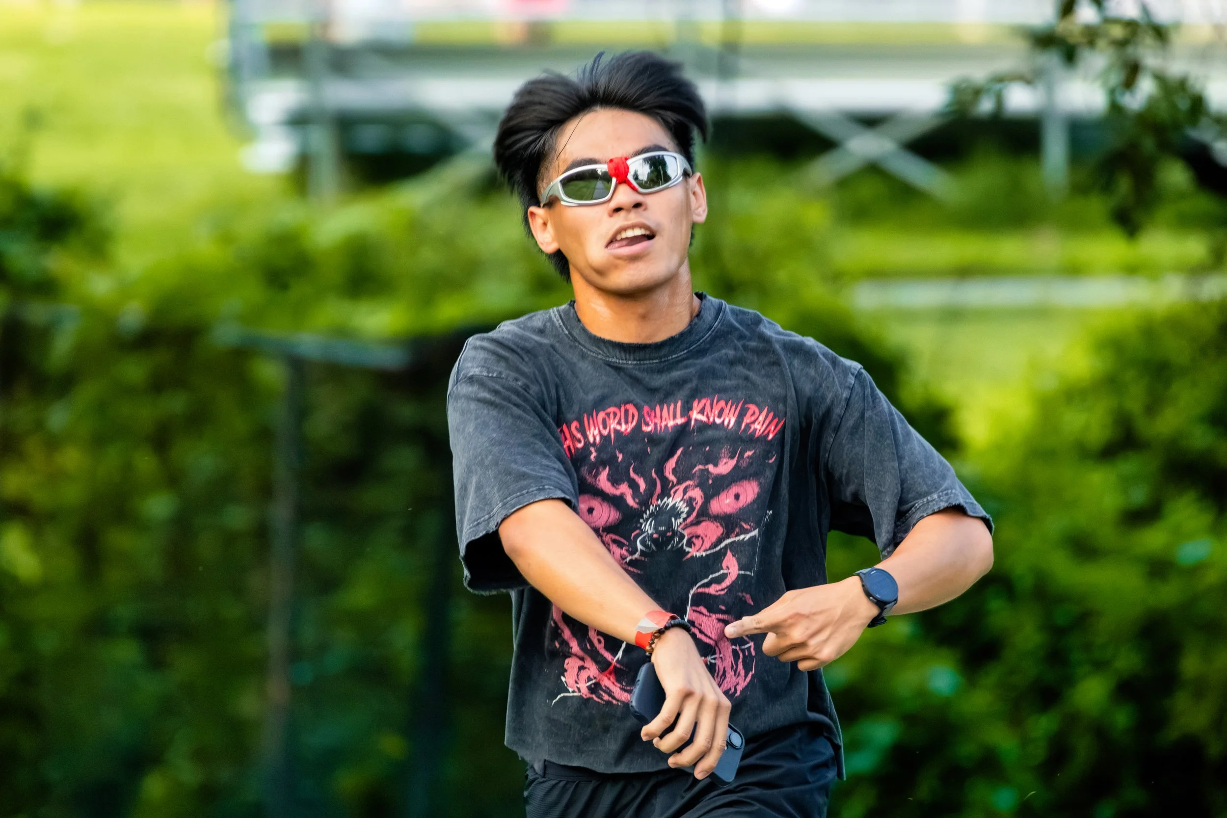 Young man with dark hair wearing reflective sunglasses with a red cloth attached at the bridge, a graphic black T-shirt, and a wristwatch, standing outdoors in front of lush green foliage.