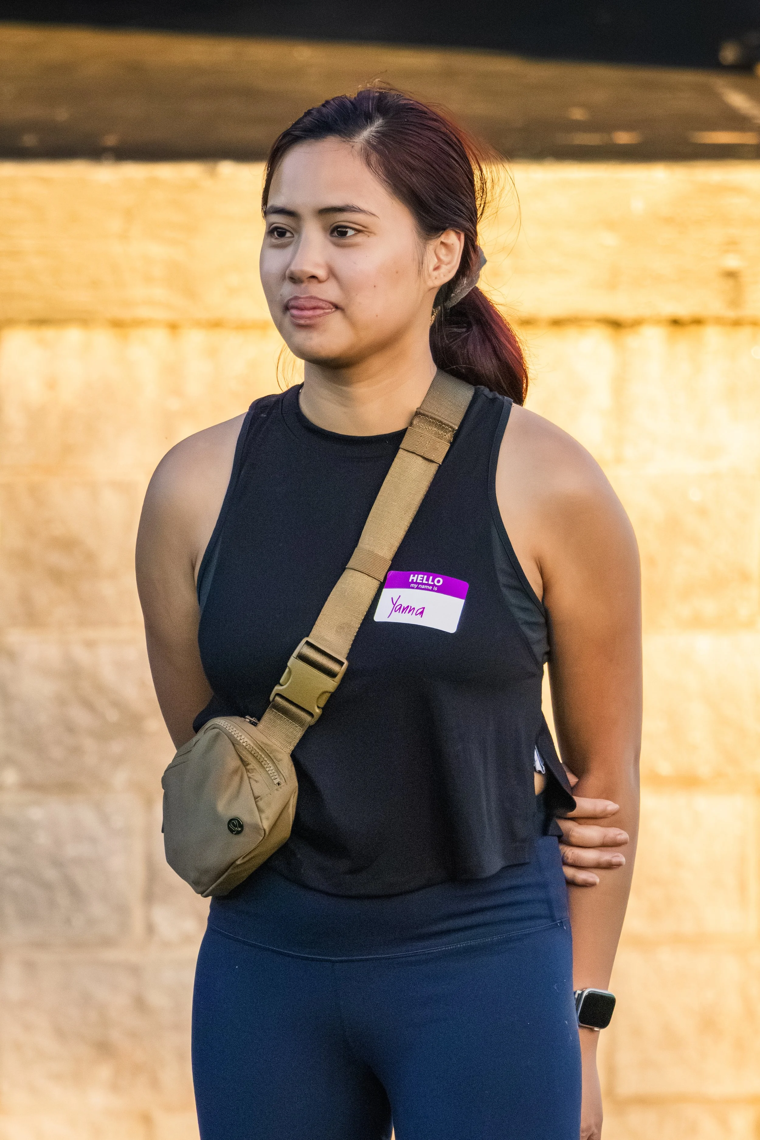 A young woman with long dark hair tied back, wearing a black sleeveless top, dark blue leggings, and a tan crossbody bag, standing outdoors near a body of water during sunset. She has a purple name tag that says "Yanna" and appears to be at an event.