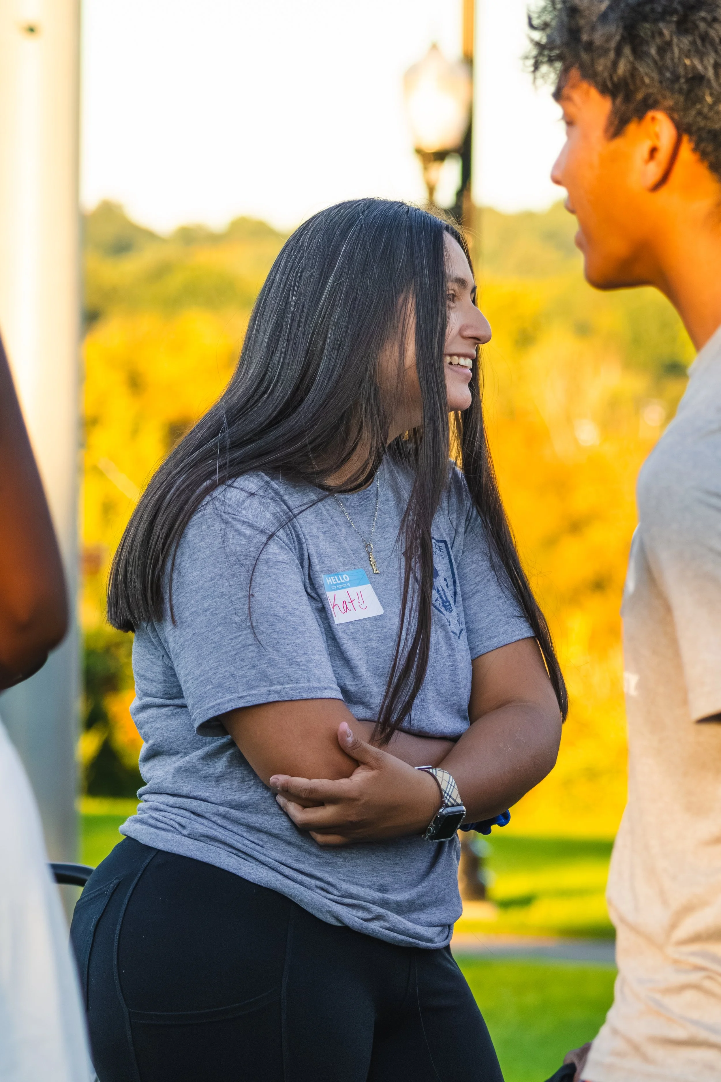 A woman with long black hair, wearing a gray t-shirt and black pants, smiling and looking at a man outside in a park during sunset.