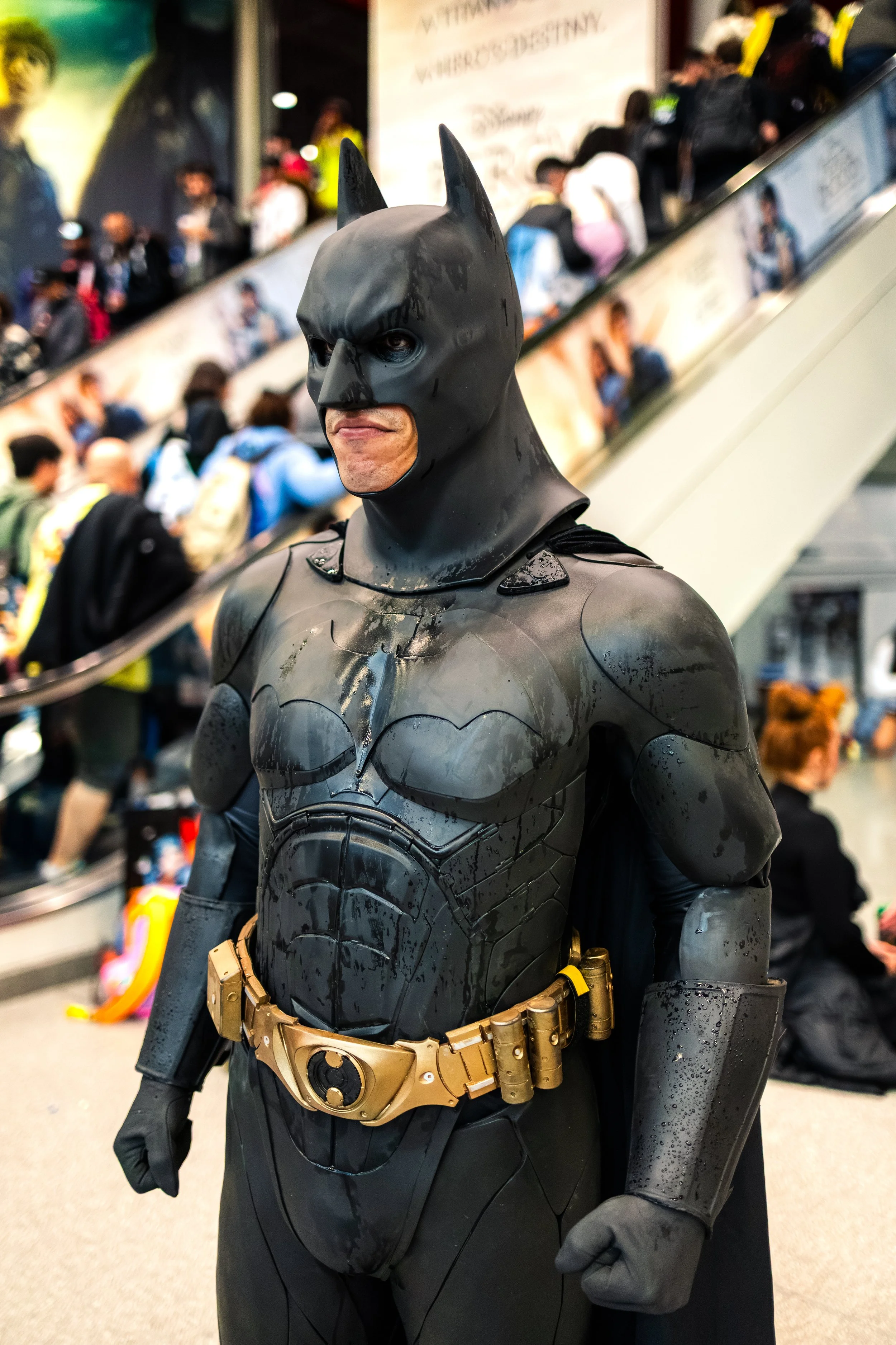 Person dressed as Batman in a costume with a black mask and cape, standing in a crowded indoor space.