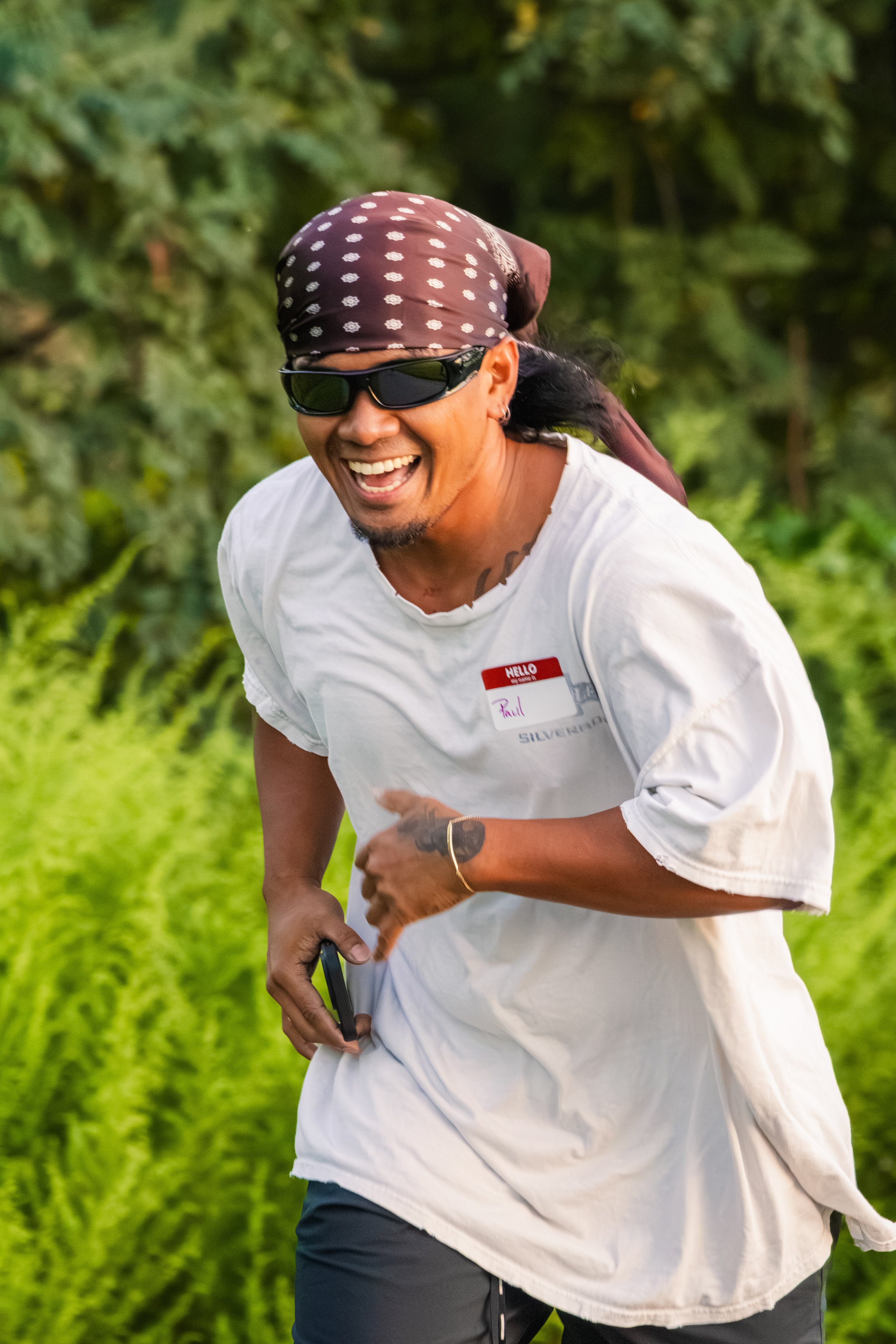A man smiling while running outdoors in a lush green area, wearing sunglasses, a bandana, a white shirt with a name tag, and black pants.