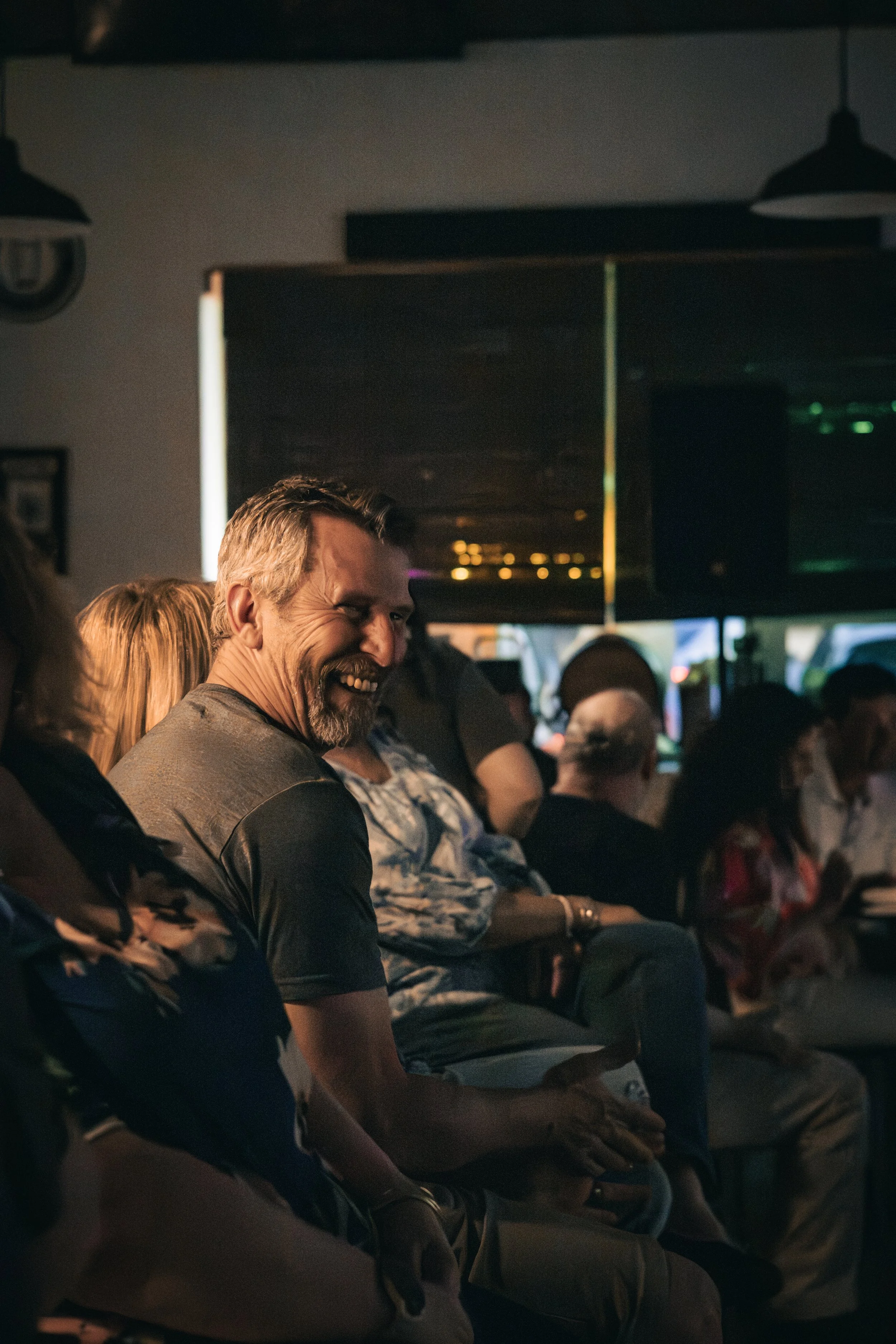 A group of people sitting in a dimly lit room, smiling and appearing to enjoy themselves. The focus is on a smiling middle-aged man with a beard and short hair, wearing a gray shirt. The background shows other seated individuals and wall decorations,