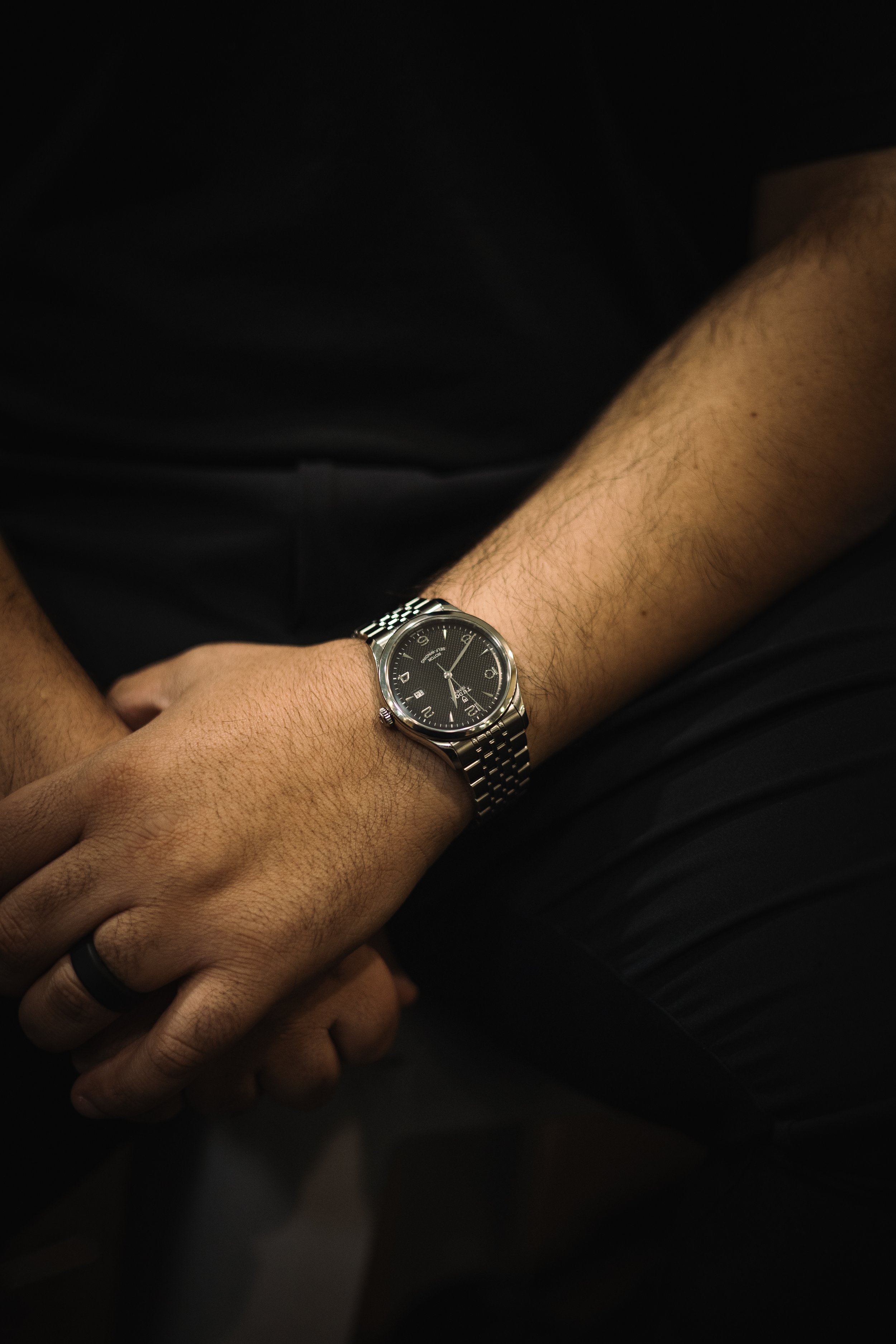 Close-up of a person wearing a silver wristwatch with a black face, resting their hand on their lap, dressed in black shorts and a black shirt.