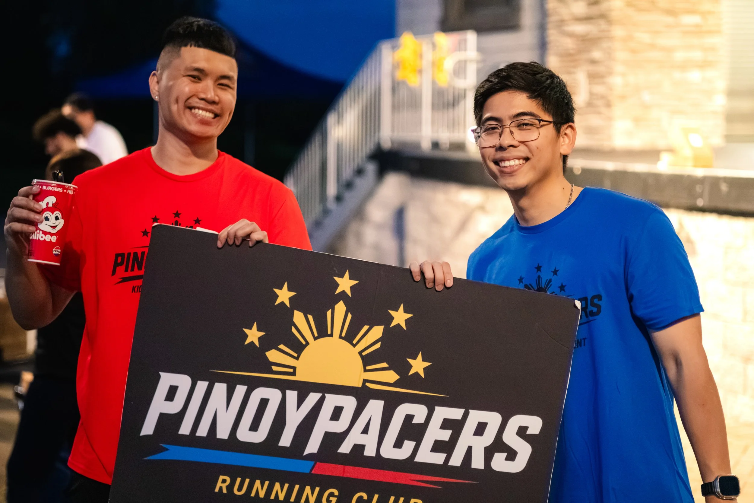 Two smiling young men, one in a red shirt holding a drink, and the other in a blue shirt, holding a sign with a sun and stars, standing outdoors at night.