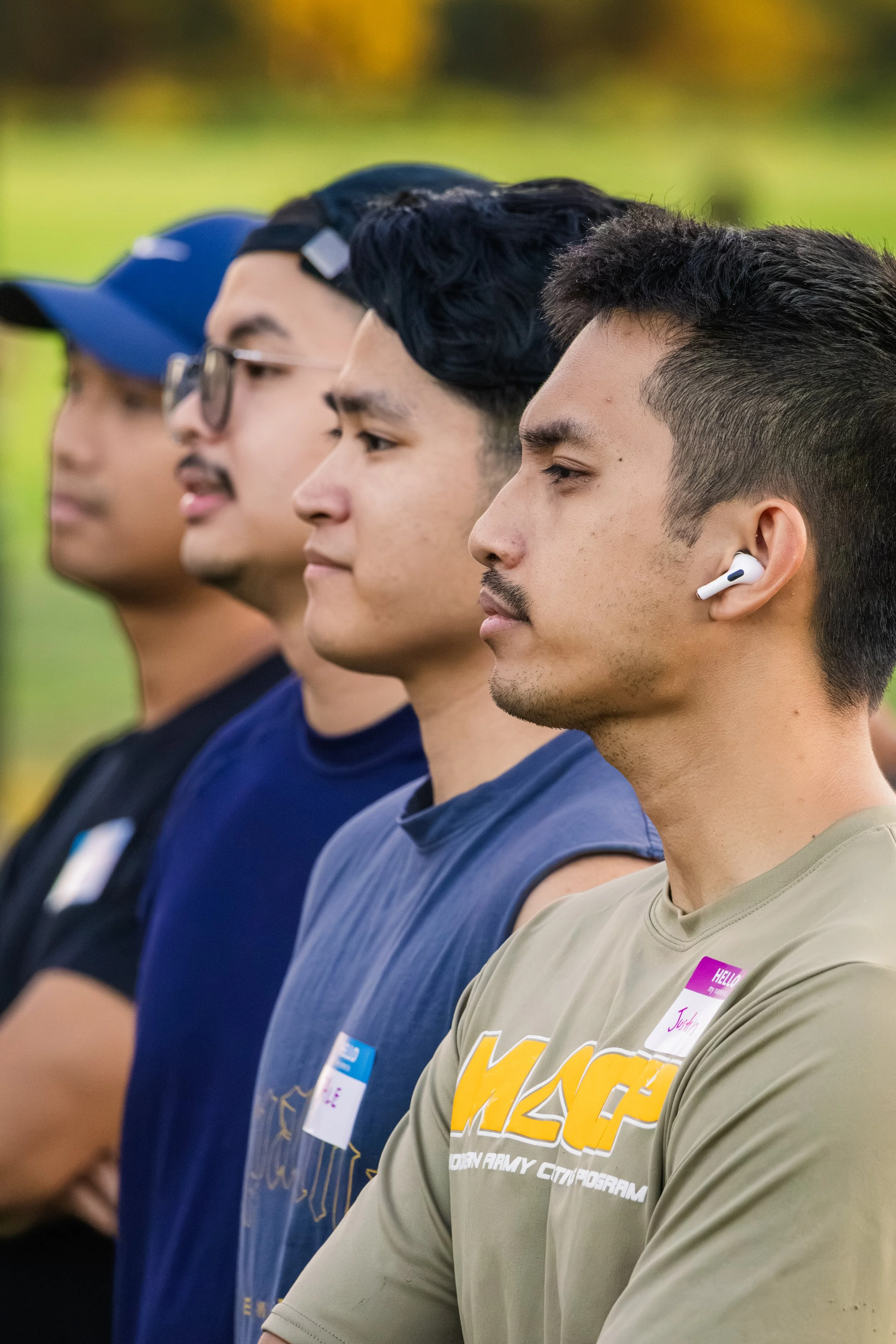 Four young men standing in a line outdoors, wearing casual shirts and t-shirts, some with name tags, appearing to be attending an event or gathering.