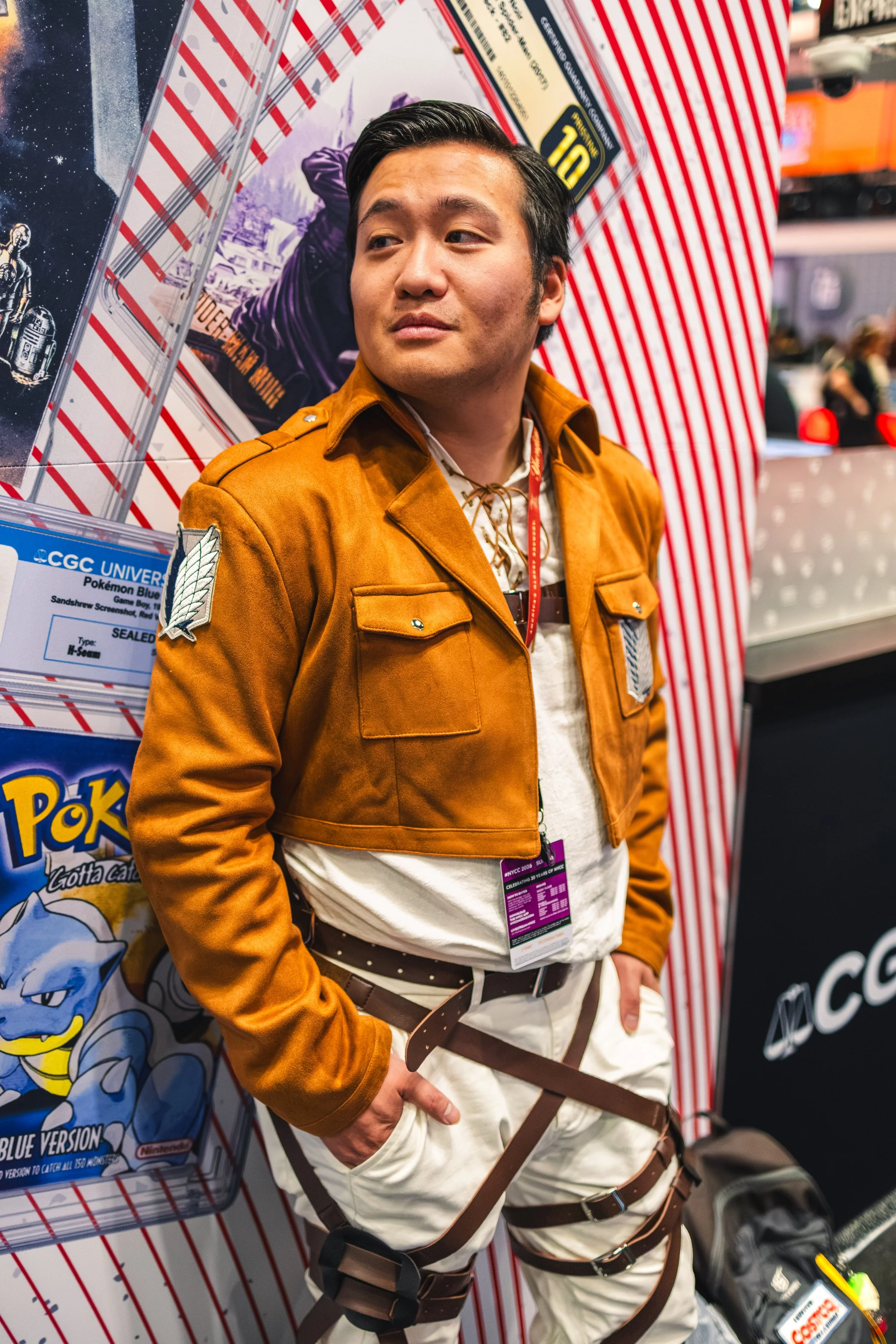 A young man with neatly combed black hair, wearing a brown jacket over a light shirt and white pants with a black belt, stands in front of a display at a convention or event. Behind him is a large poster featuring Pokémon characters, including a blue
