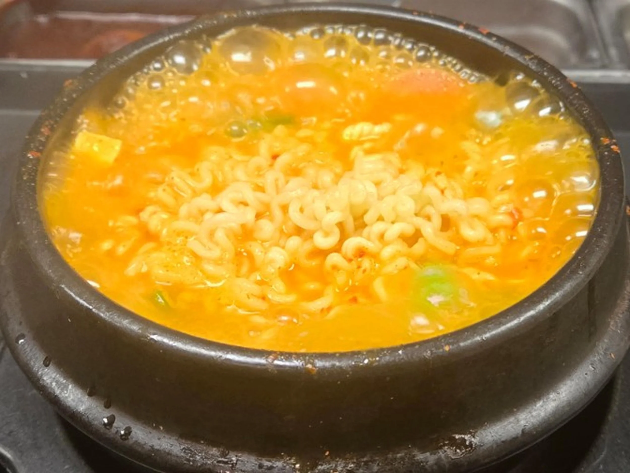 Cooked instant ramen noodles boiling in a black pot, with some broth and small pieces of green vegetables visible.