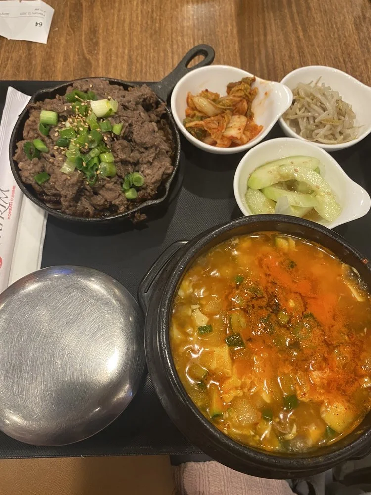 A Korean meal on a black tray featuring a hot stone bowl of beef with scallions, a bowl of spicy soup with vegetables, and three small dishes of kimchi, bean sprouts, and pickled cucumbers.