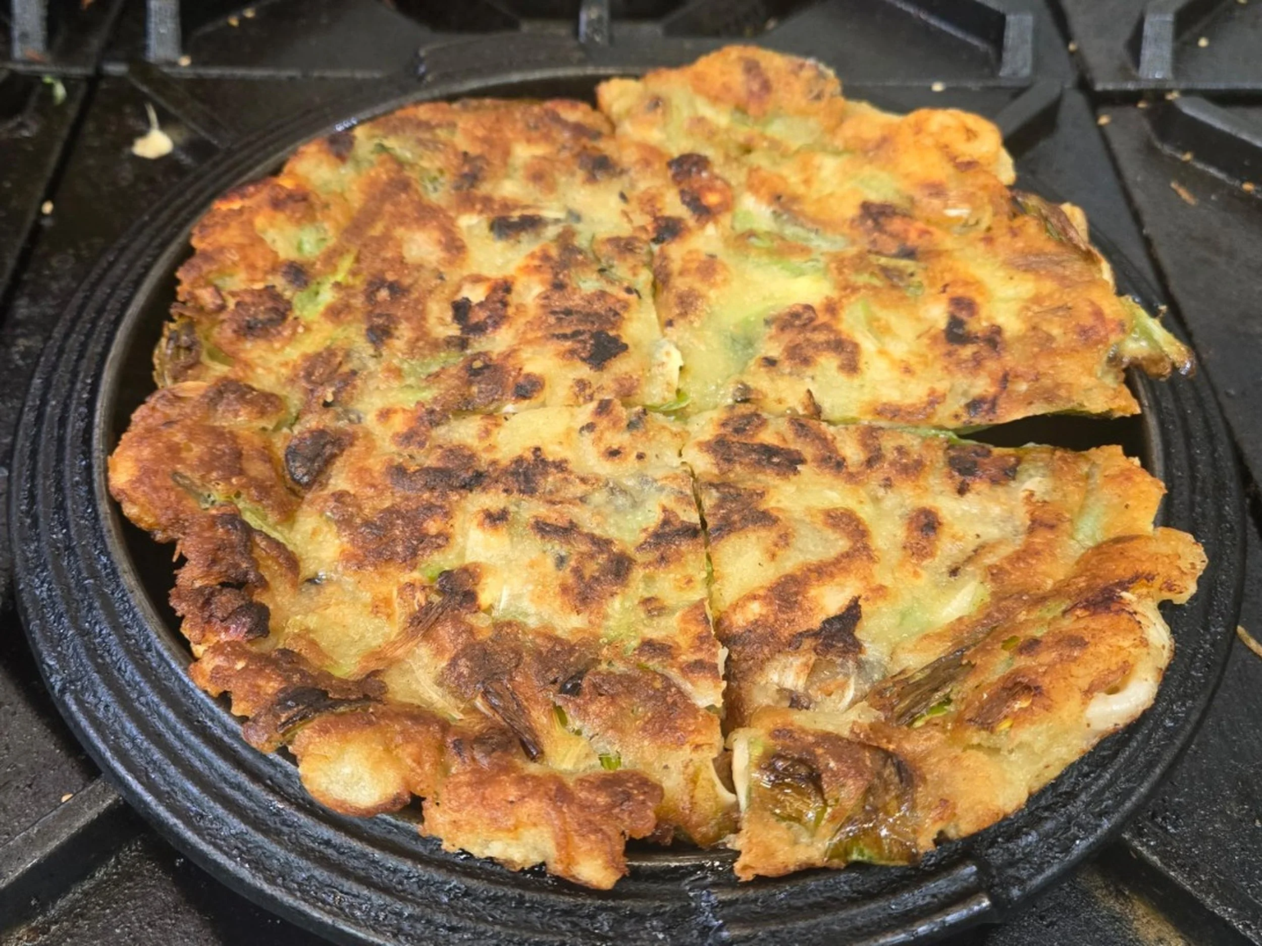 A close-up of a cooked vegetable pancake with green vegetables and crispy, golden-brown edges, baking in a black cast iron skillet.