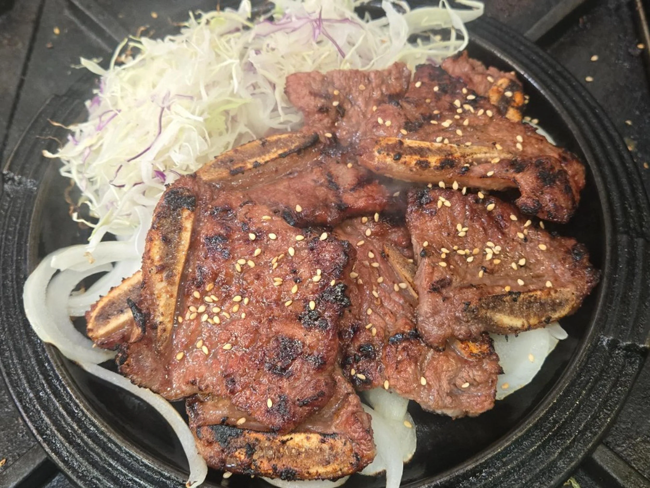 Grilled beef steaks topped with sesame seeds on a black grill, garnished with sliced onions and shredded cabbage on the side.