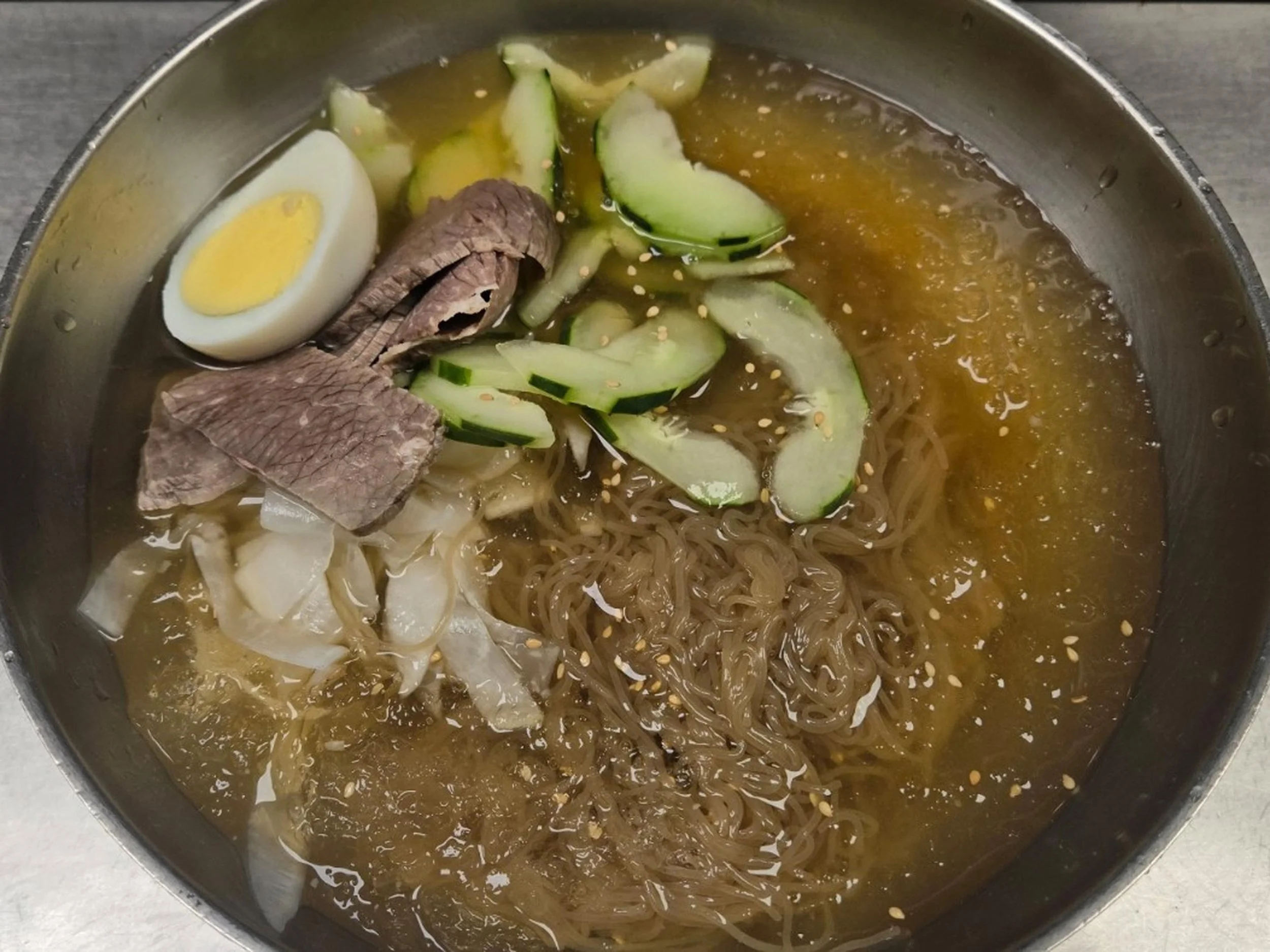 A pot of ramen noodles with sliced cucumbers, half a boiled egg, and slices of beef, garnished with sesame seeds and a soy-based broth.
