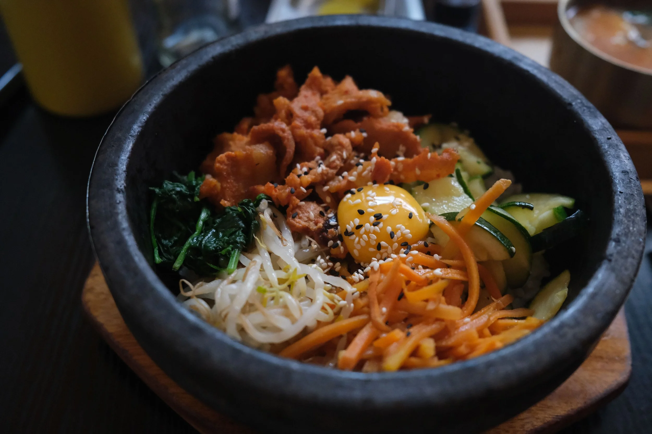 A black stone bowl filled with a Korean dish called bibimbap, including rice, sautéed spinach, shredded carrots, zucchini, bean sprouts, a fried egg, tender marinated beef, and sprinkled sesame seeds.