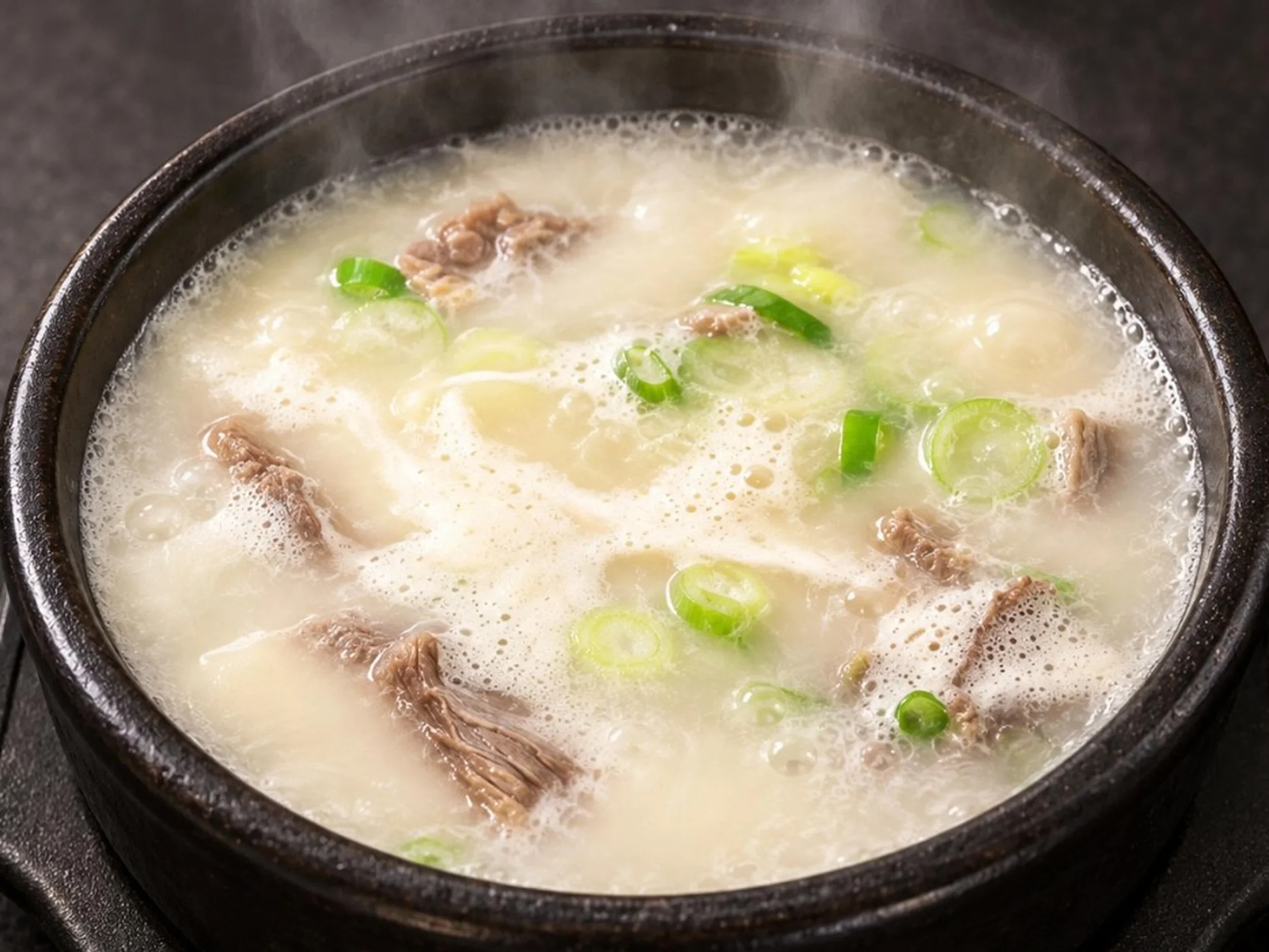 A pot of Korean stew with slices of green onions, shredded meat, and a milky broth.