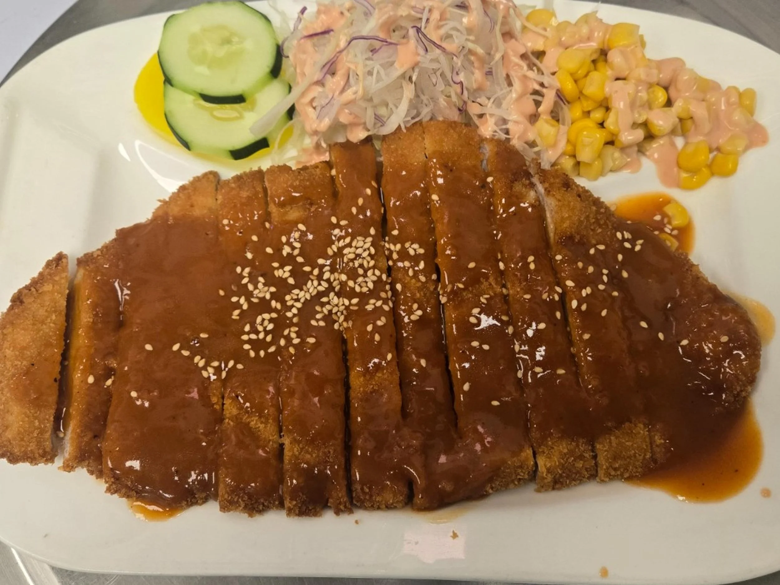 Breaded fried cutlet topped with brown sauce and sesame seeds, served with sliced cucumber, coleslaw, and corn salad on a white plate.