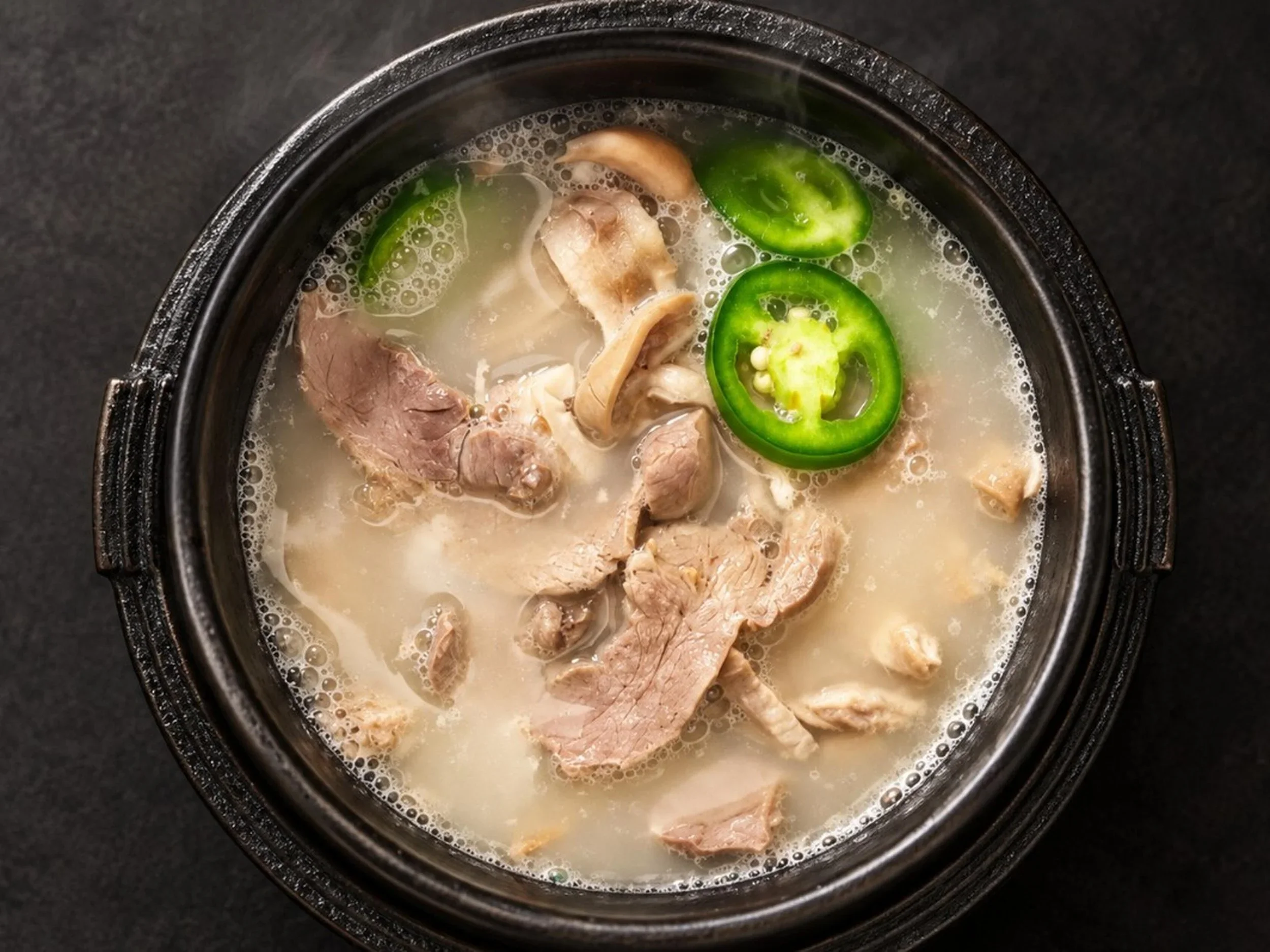 A black pot containing hot soup with pieces of pork, slices of jalapeño pepper, and possibly mushrooms in a light broth.