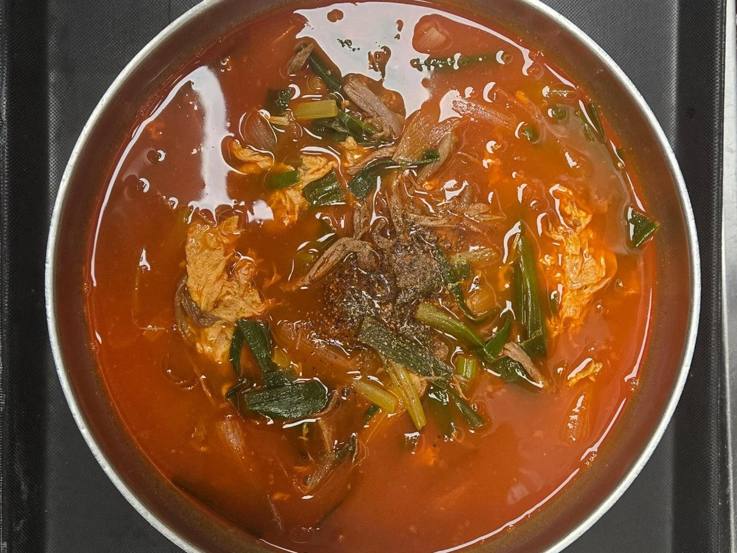 A pot of simmering stew with chunks of meat, green vegetables, and a red broth, sitting on a black stove.