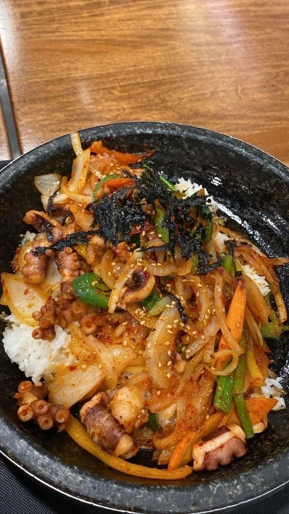 Bowl of Asian stir-fry with vegetables, octopus, rice, and seaweed, served in a black bowl on a wooden table.