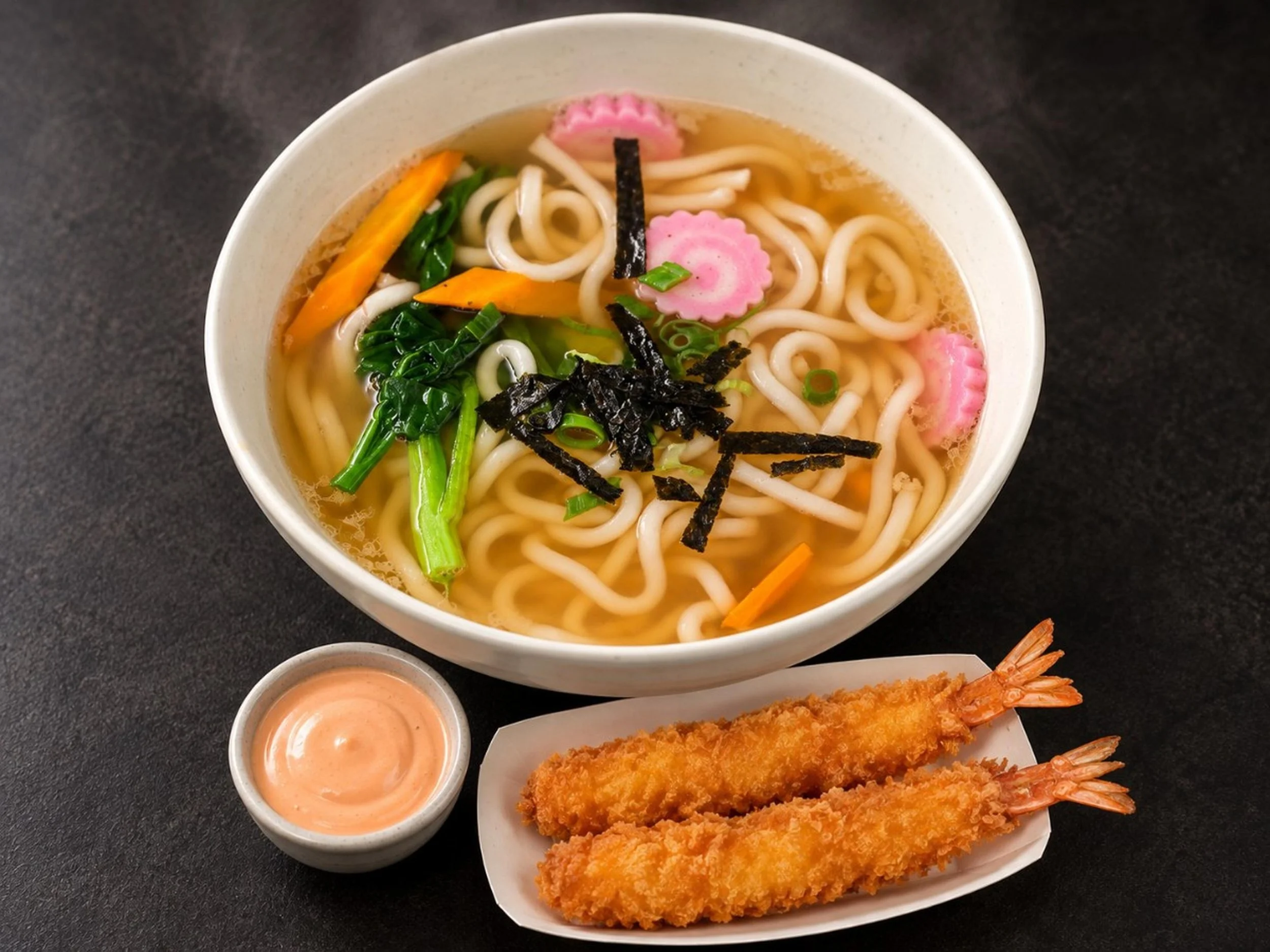A bowl of ramen with sliced pink fish cake, seaweed strips, green onions, carrots, and noodles in broth. A plate of fried shrimp tempura and a small bowl of dipping sauce are partially visible at the bottom.