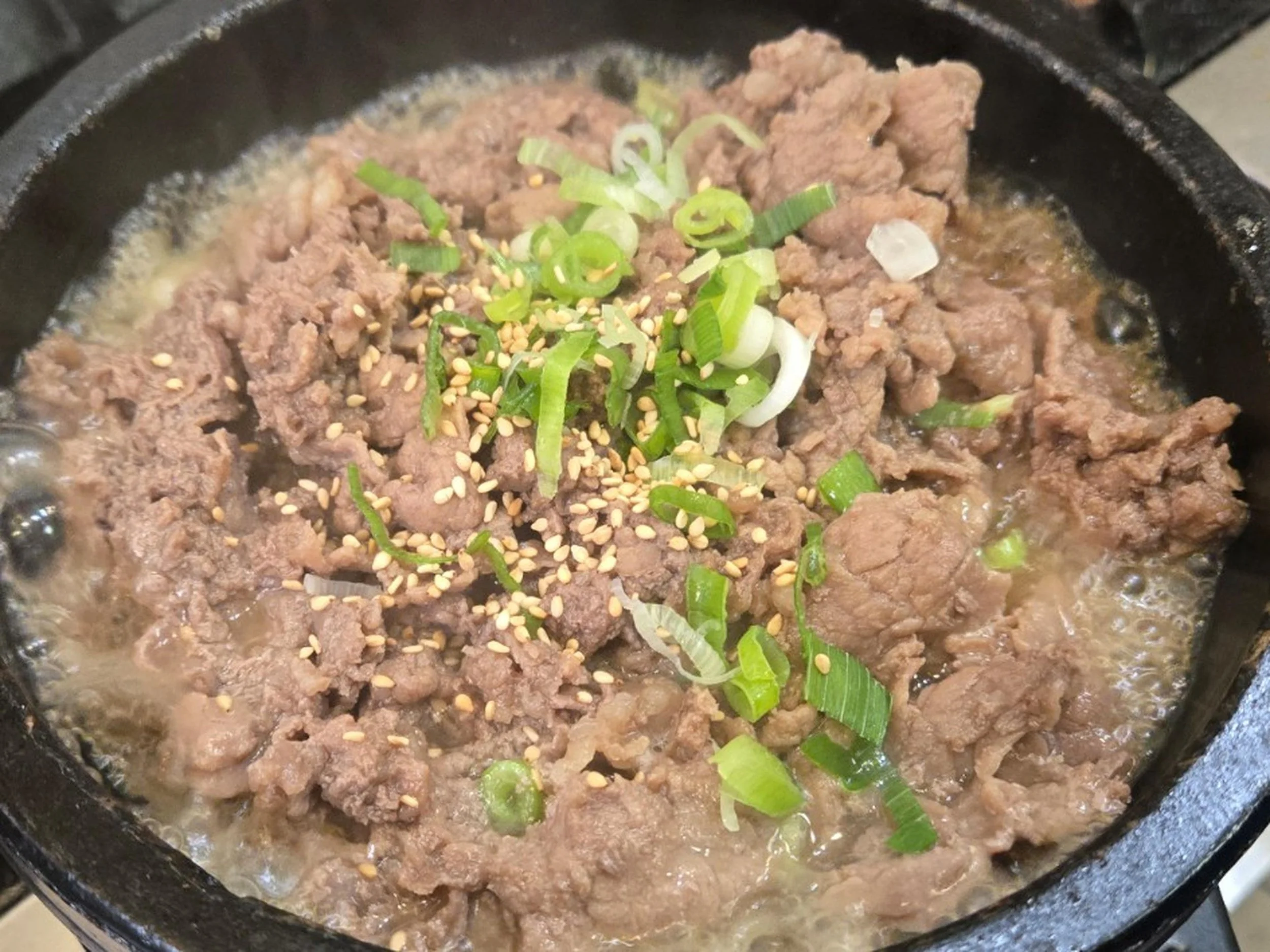 Ground beef in a skillet garnished with chopped green onions and sesame seeds.