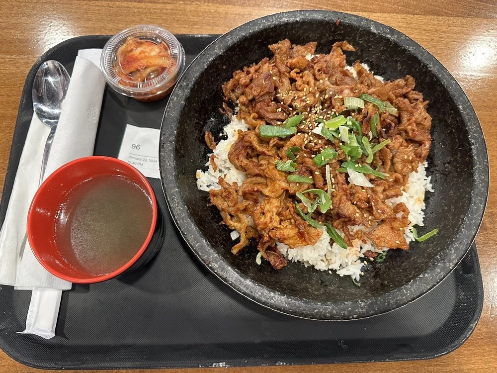 A meal served on a black tray featuring a hot stone bowl with rice and stir-fried beef topped with green onions, a small bowl of soup, a small container of kimchi, a spoon, and a napkin.