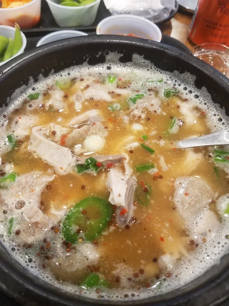 A hot pot of broth with slices of green jalapeño, small red chili flakes, bits of chicken, and green onions.