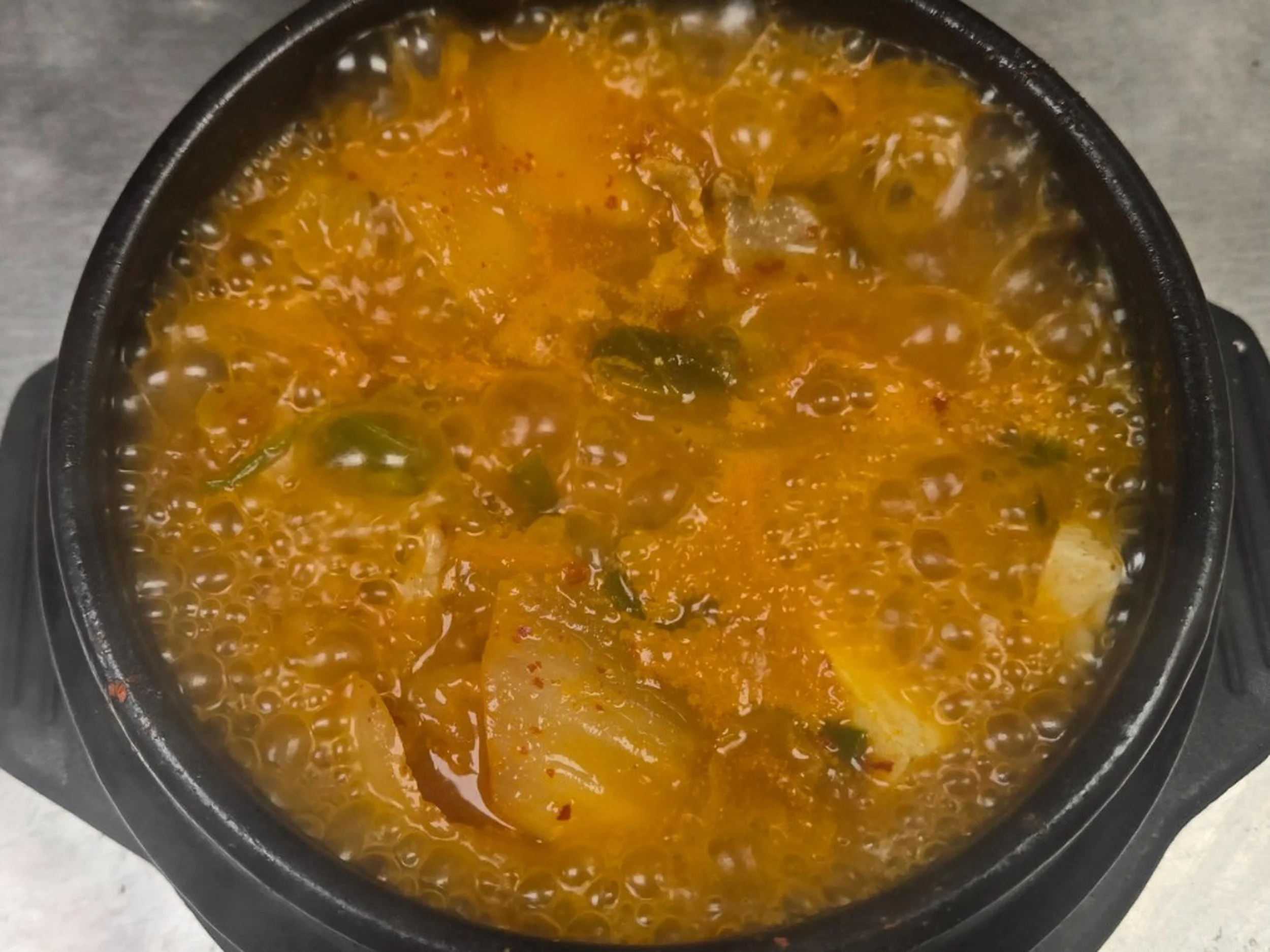 A bowl of hot, spicy soup with vegetables and herbs, seen from above on a gray surface.