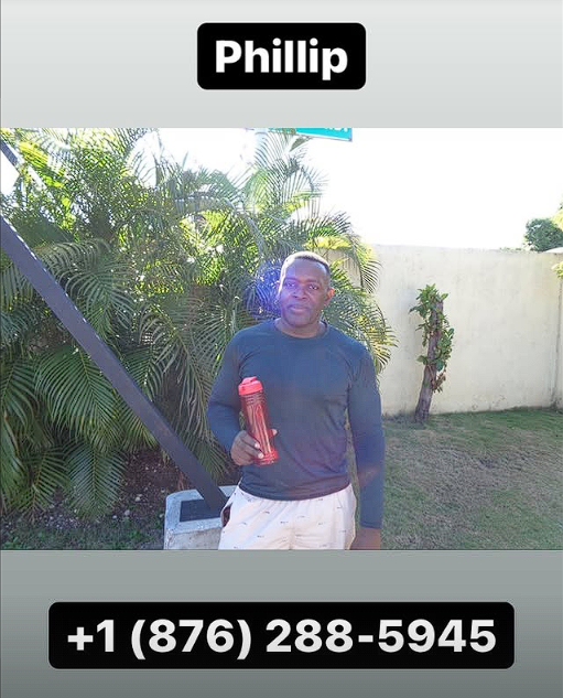A man standing outdoors in front of green palm plants, wearing a navy blue long-sleeve shirt and beige pants, holding a red water bottle, with a white wall and grass in the background. The image has his name, Phillip, and a phone number, +1 (876) 288-5945, displayed at the top and bottom.