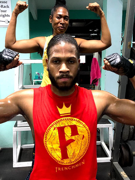 A man in a red tank top flexing his muscles and a woman in a yellow top behind him, also flexing her muscles, in a gym setting.