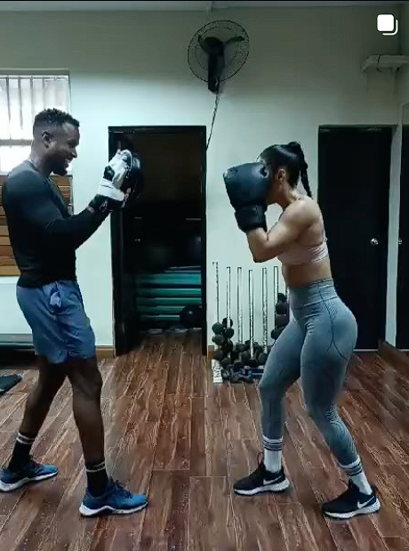 A man and a woman are boxing in a gym. The man is smiling and throwing a punch, while the woman is wearing boxing gloves and preparing to punch back. They are standing on a wooden floor with gym equipment in the background.