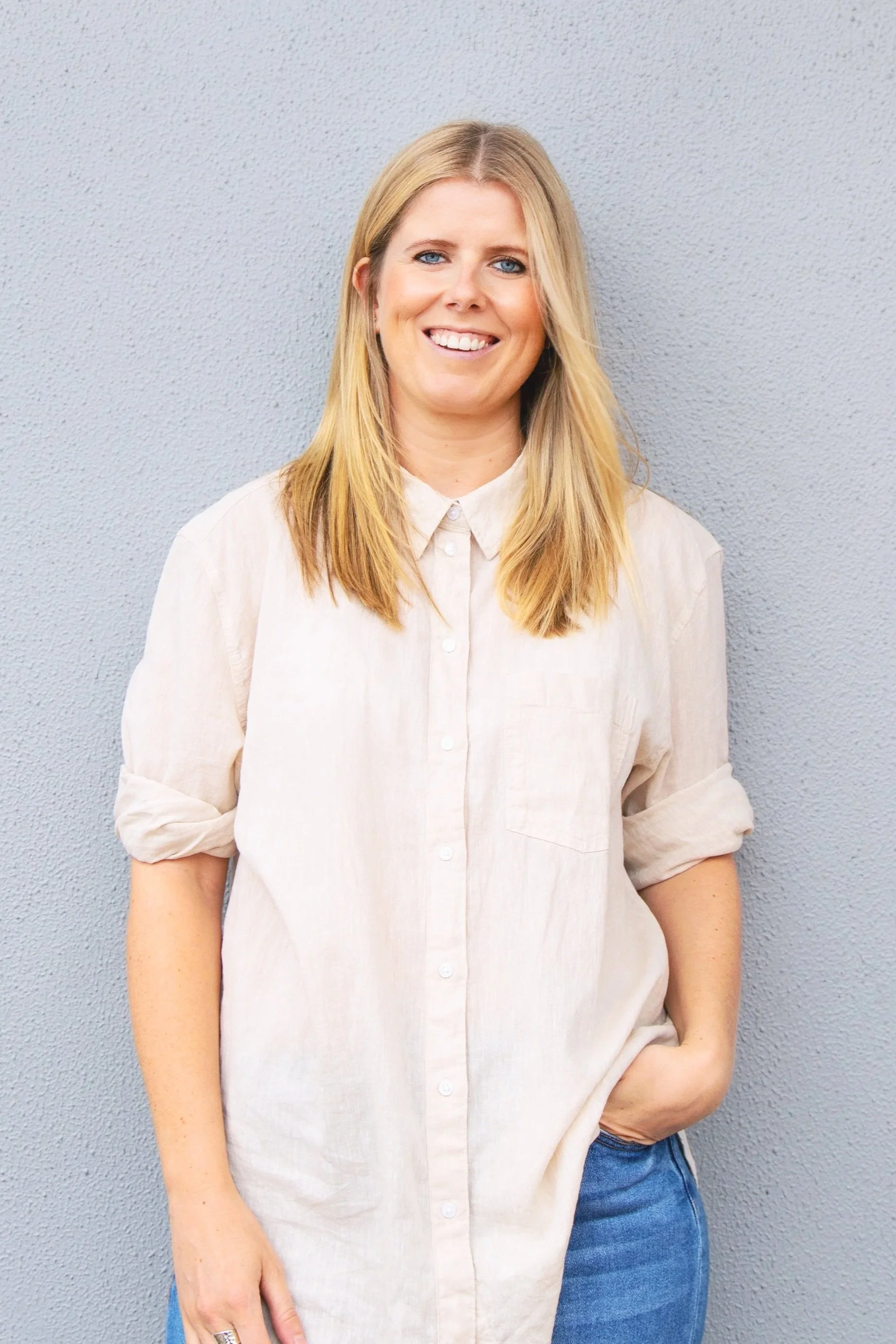 Woman with blonde hair wearing a light beige shirt and blue jeans standing against a gray textured wall, smiling and looking at the camera.