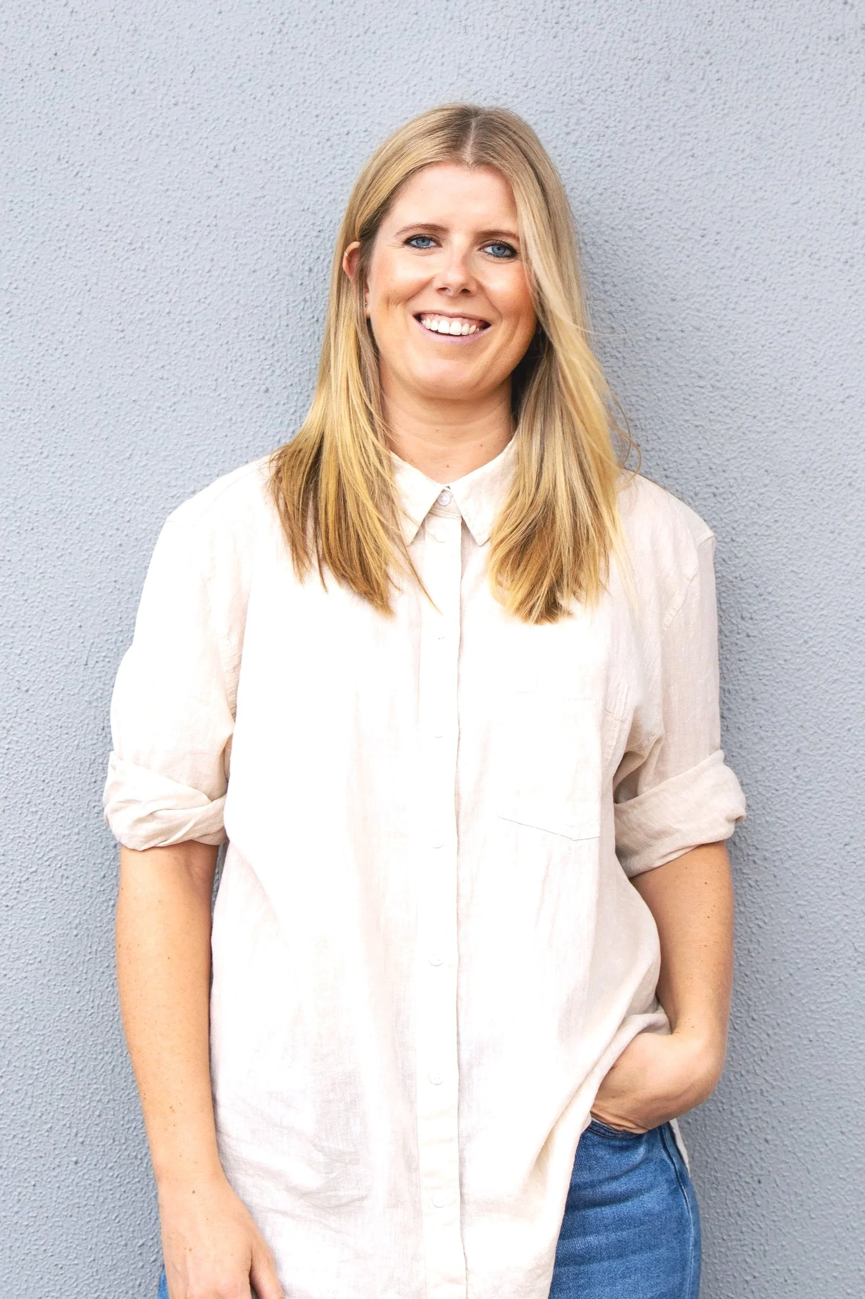 A woman with blonde hair, wearing a cream-colored shirt with rolled-up sleeves, standing against a gray textured wall, smiling with her teeth showing and looking at the camera.