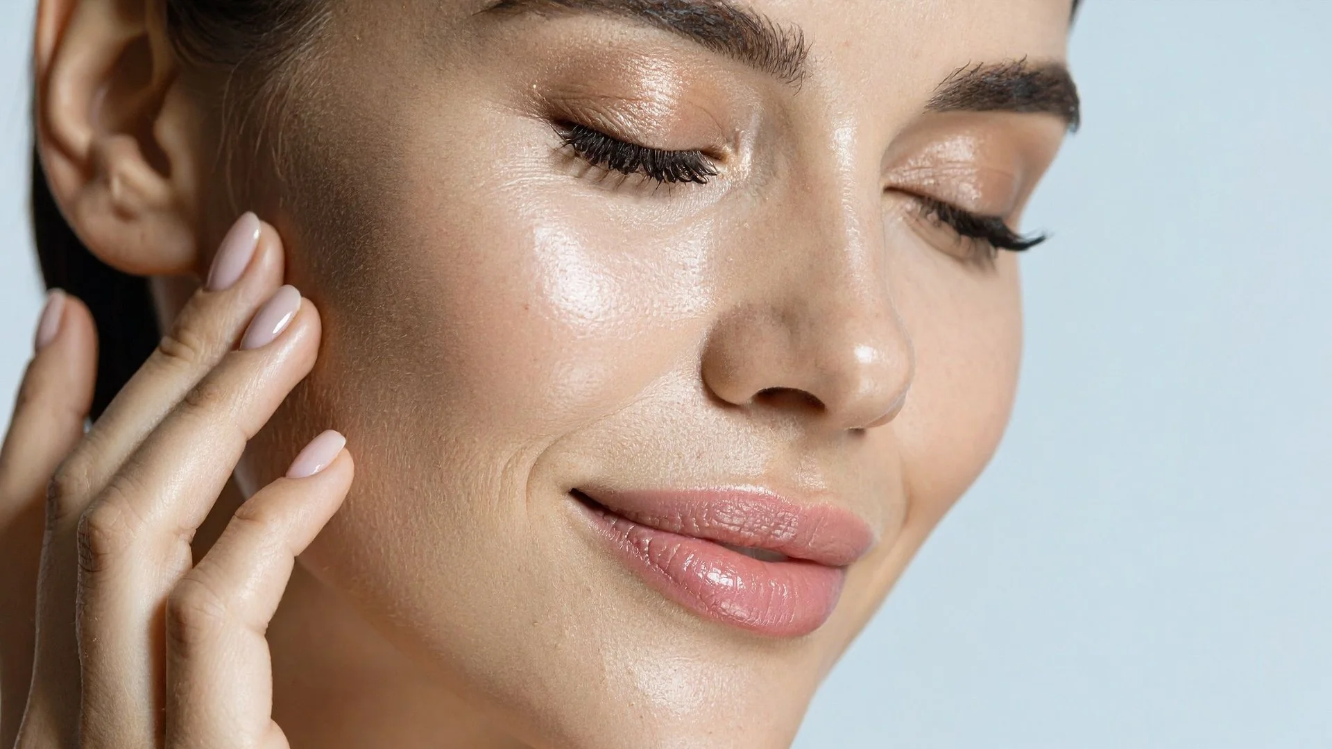 Close-up of a woman with smooth, glowing skin, touching her face with her fingers, wearing natural makeup and closed eyes.