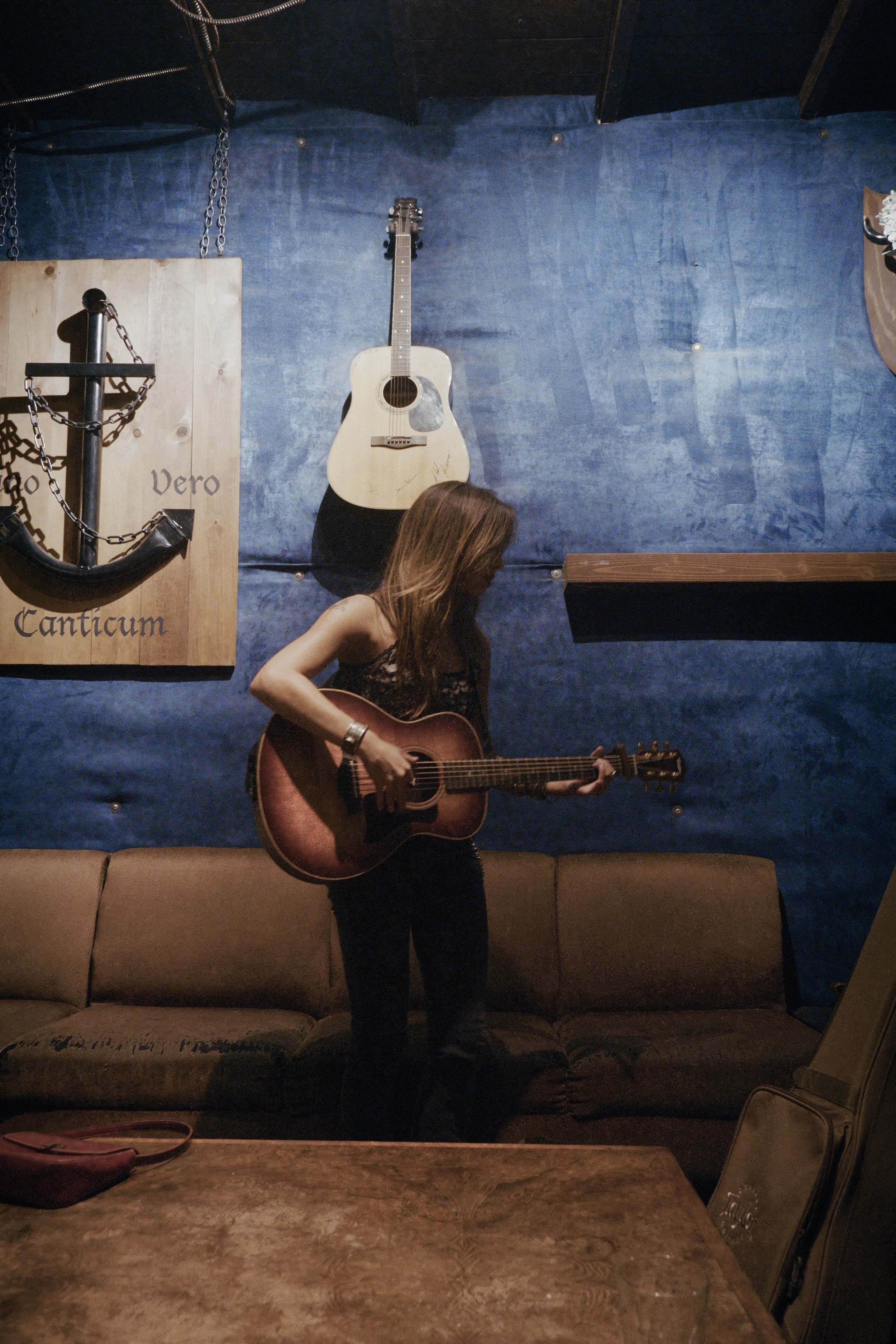 A woman with long brown hair playing an acoustic guitar, standing in a dimly lit room with a blue wall, a beige couch, and musical decorations including a large anchor with chains and a guitar hanging on the wall.