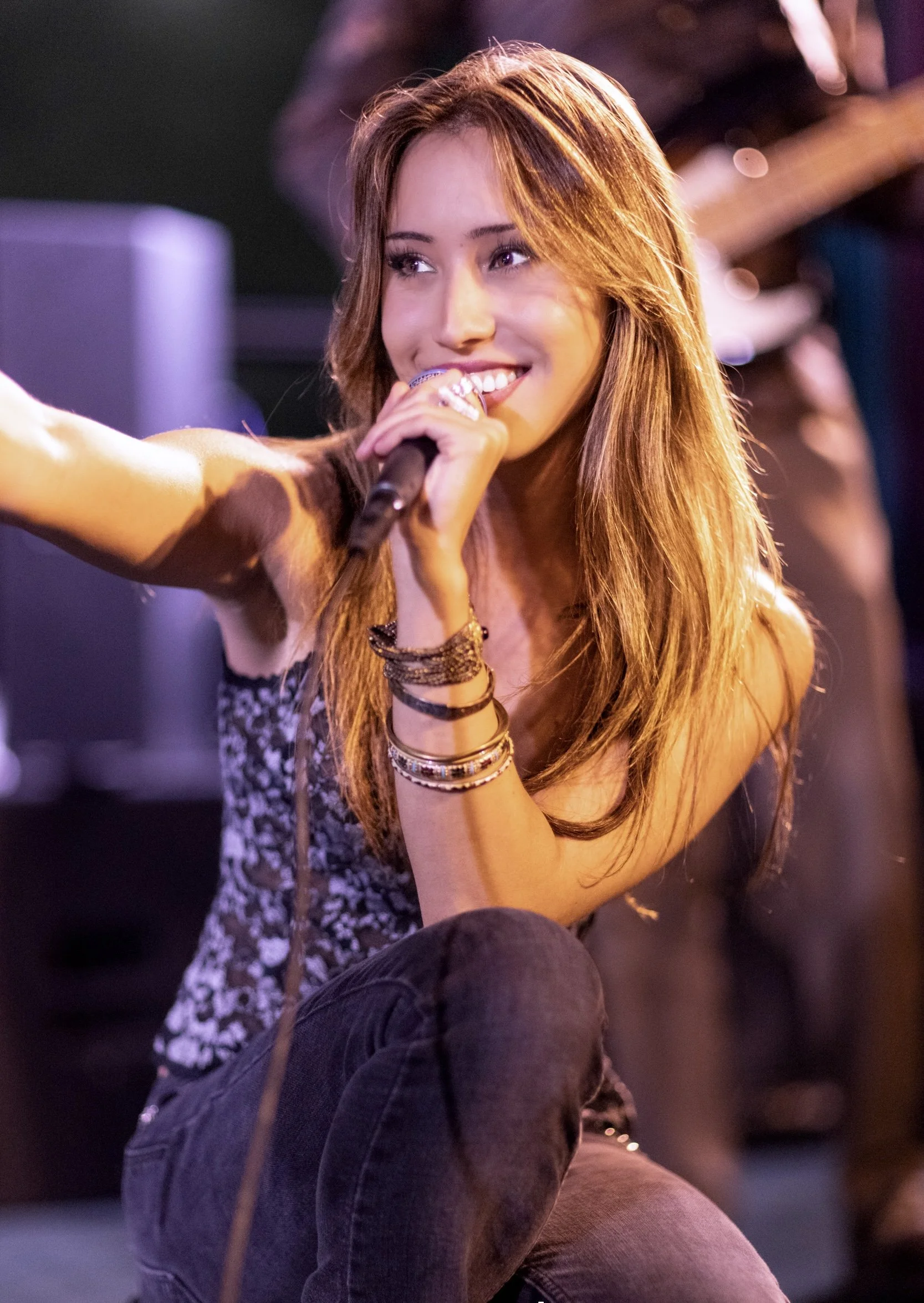 A young woman with long reddish-brown hair holding a microphone and smiling, sitting with her knees up in a dimly lit setting, possibly performing or speaking.