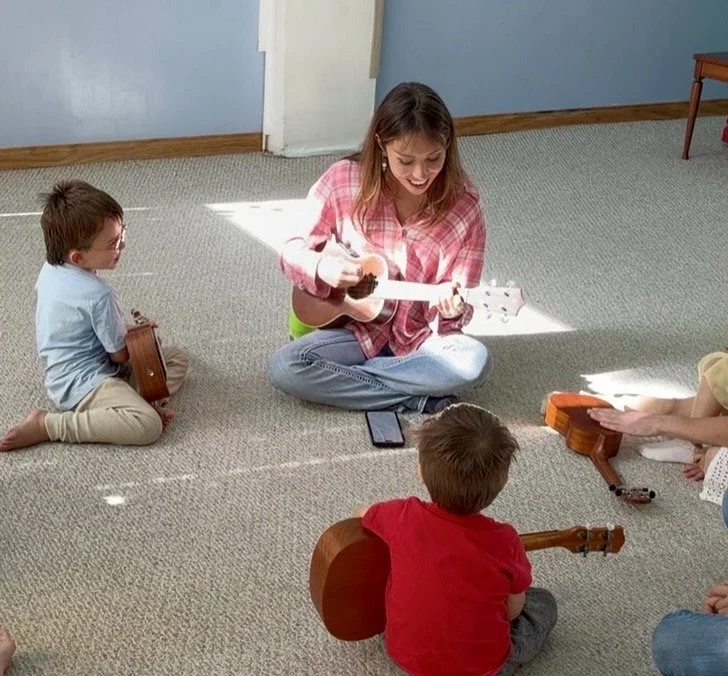 A woman and three children sitting on a carpeted floor, playing stringed instruments together in a room with light blue walls.