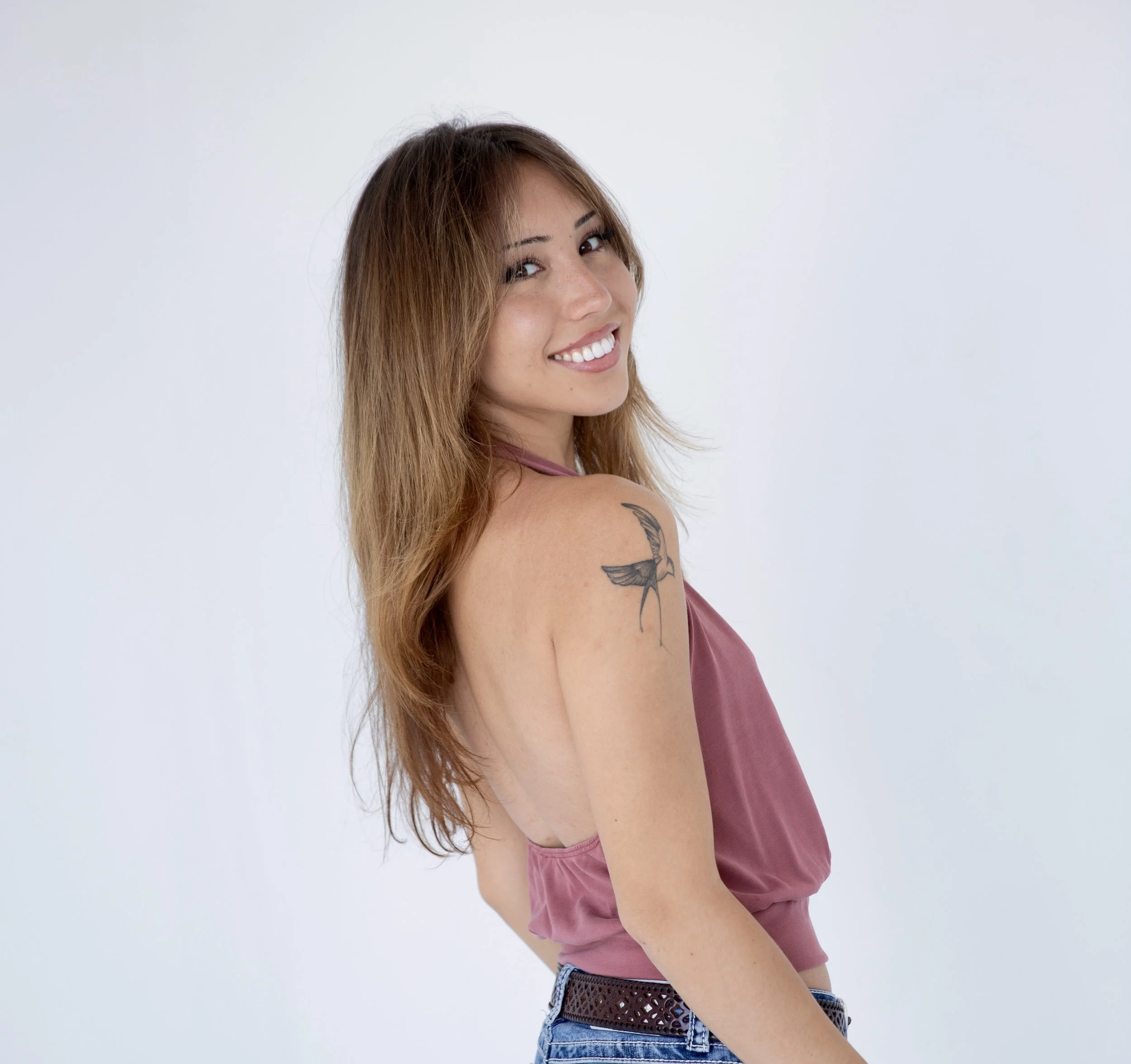 Front and side view of a smiling woman with long, light brown hair, wearing a sleeveless pink top and blue jeans, standing against a plain white background.