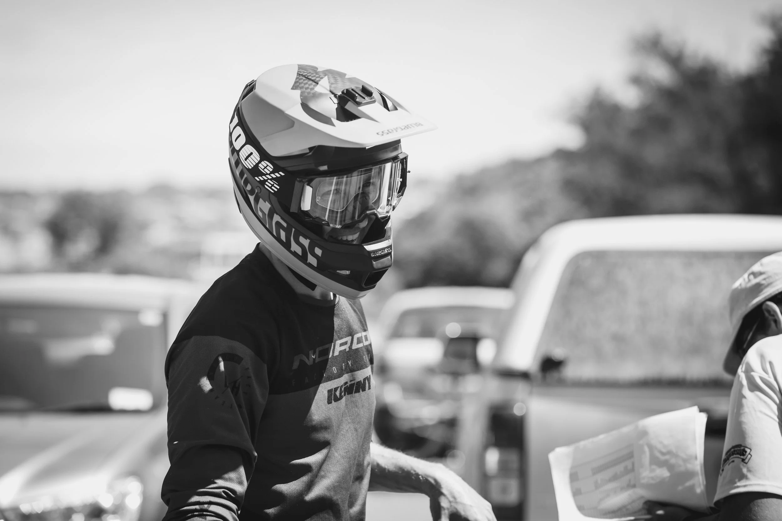 A person wearing a full-face helmet smiling, standing outdoors with parked cars in the background.