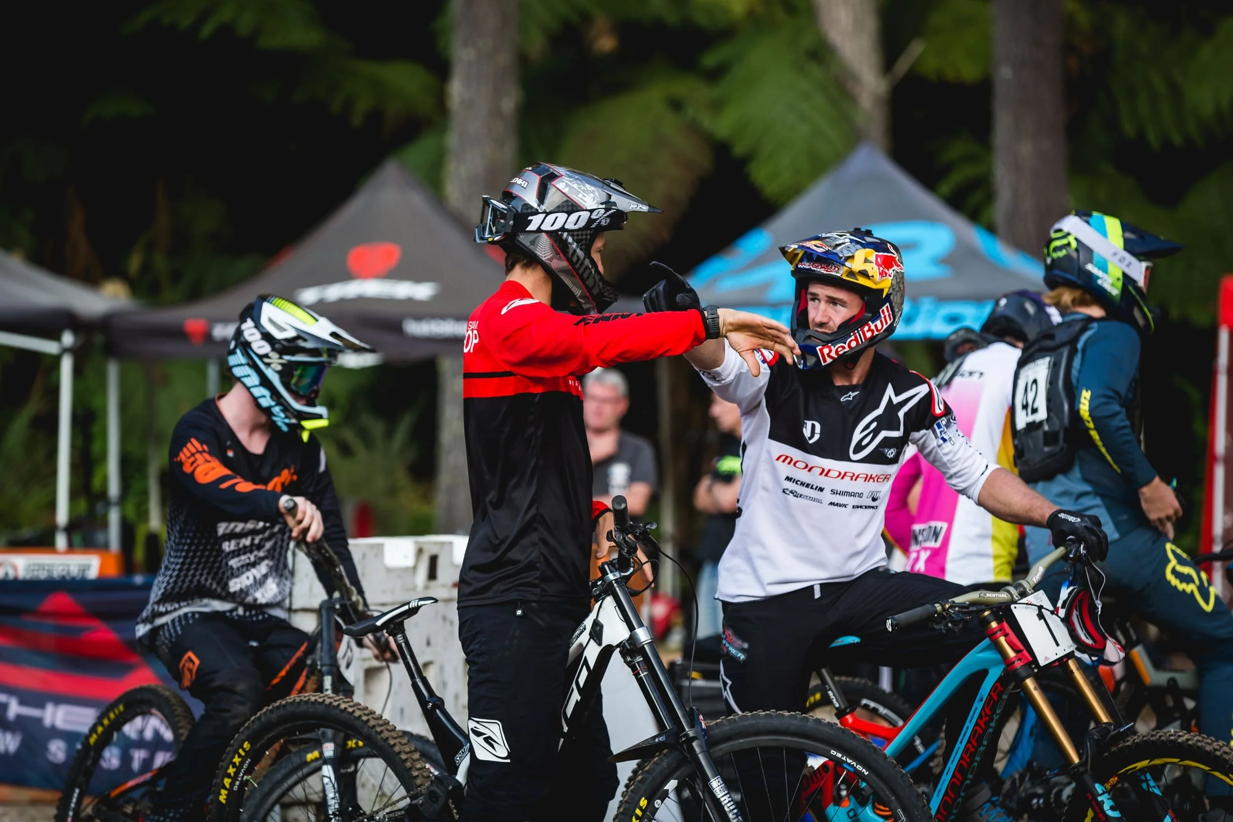 Motocross racers and team members at a race event, with some riders wearing helmets and jerseys in various colors, standing next to their bikes and engaging in conversation.