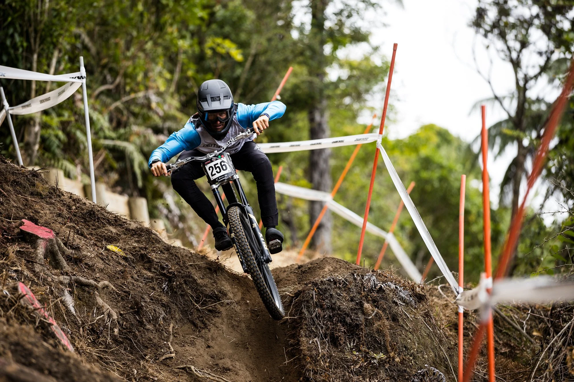 Mountain biker wearing helmet and goggles navigating a steep, muddy downhill trail marked with orange and white poles and tape, surrounded by trees.