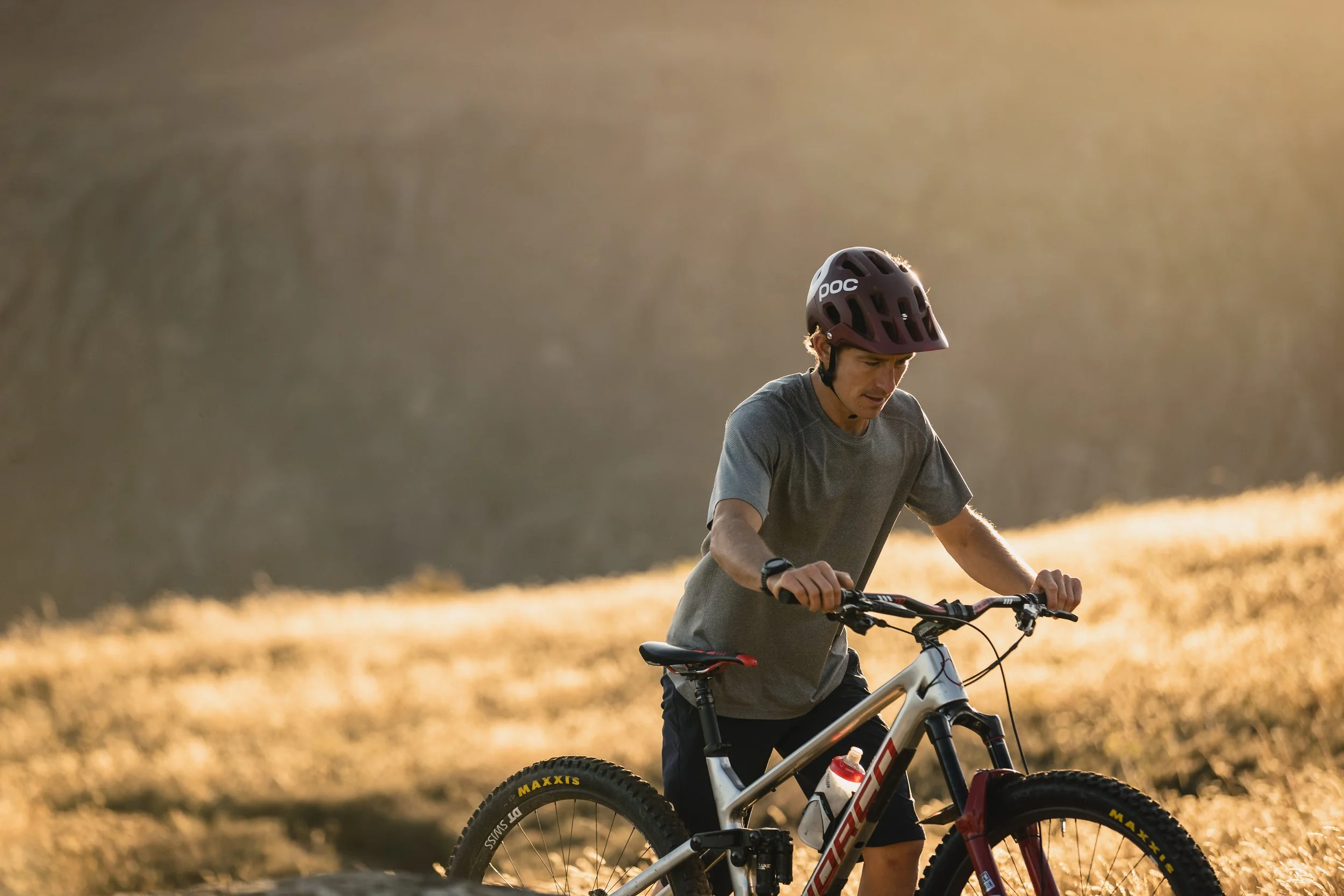 A man wearing a POC helmet riding a mountain bike through a grassy outdoor area during sunset.